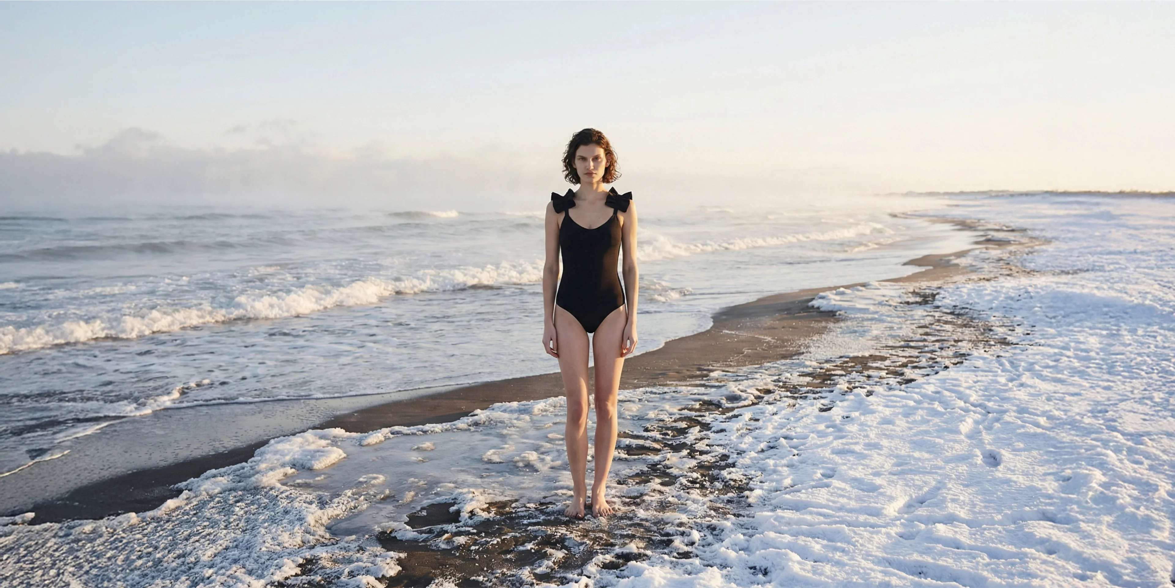 Frau im schwarzen Baeanzug steht im Winter am Meer.