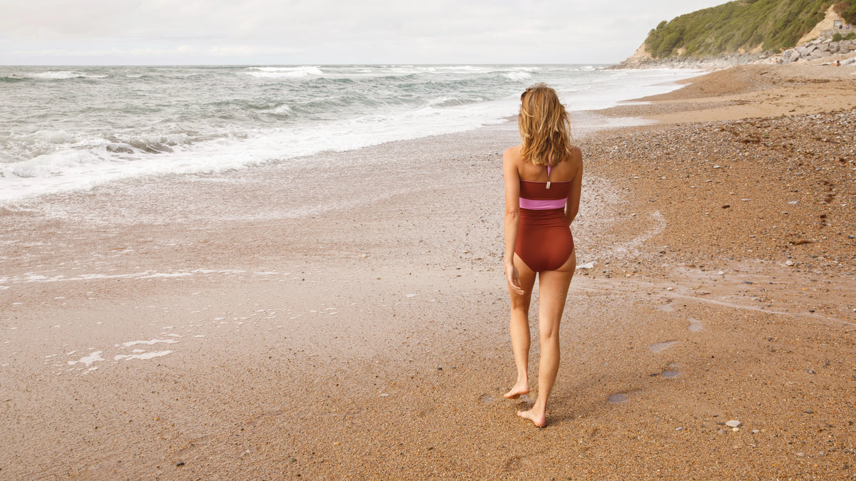 Back view of a woman walking along a pebble beach in a rust MYMARINI bandeau swimsuit with a pink stripe