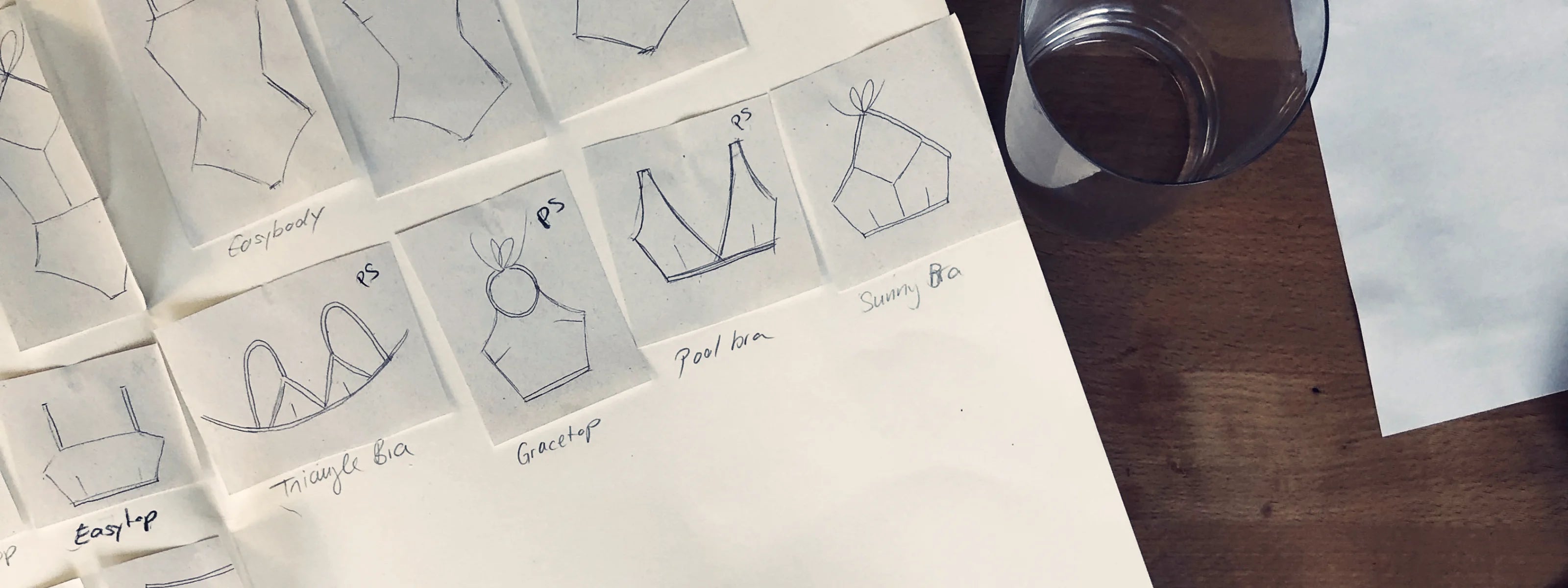 Skizzen von MYMARINI-Bikinitops auf Papier neben einem Glas Wasser, darunter Bandeau und Neckholder-Designs.