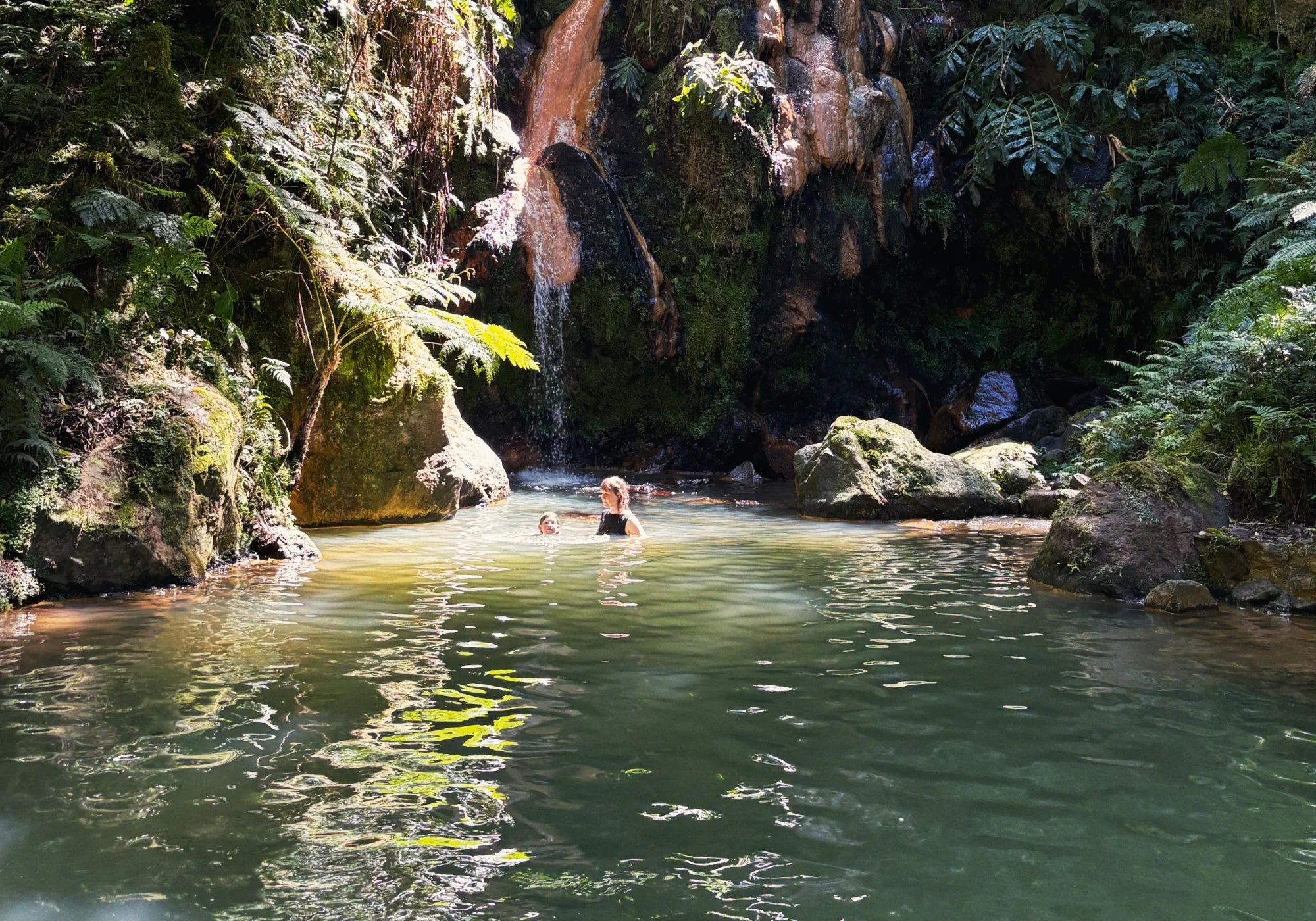 Person schwimmt in einem natürlichen Pool mit Wasserfall, umgeben von üppigem Grün.