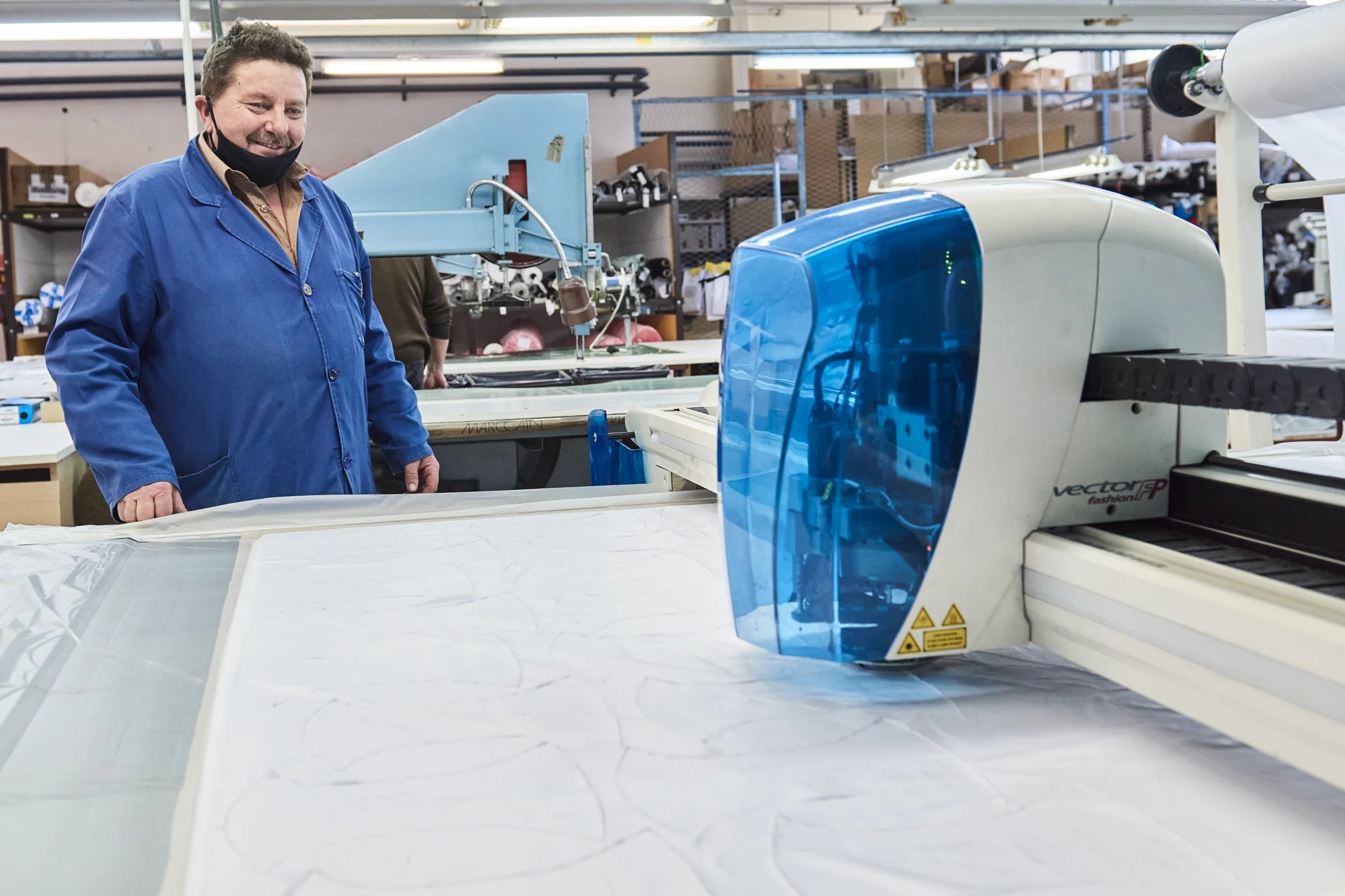 Mitarbeiter in blauer Arbeitskleidung steht neben einer automatisierten Zuschneidemaschine mit blauer Abdeckung in einer Textilfabrik.