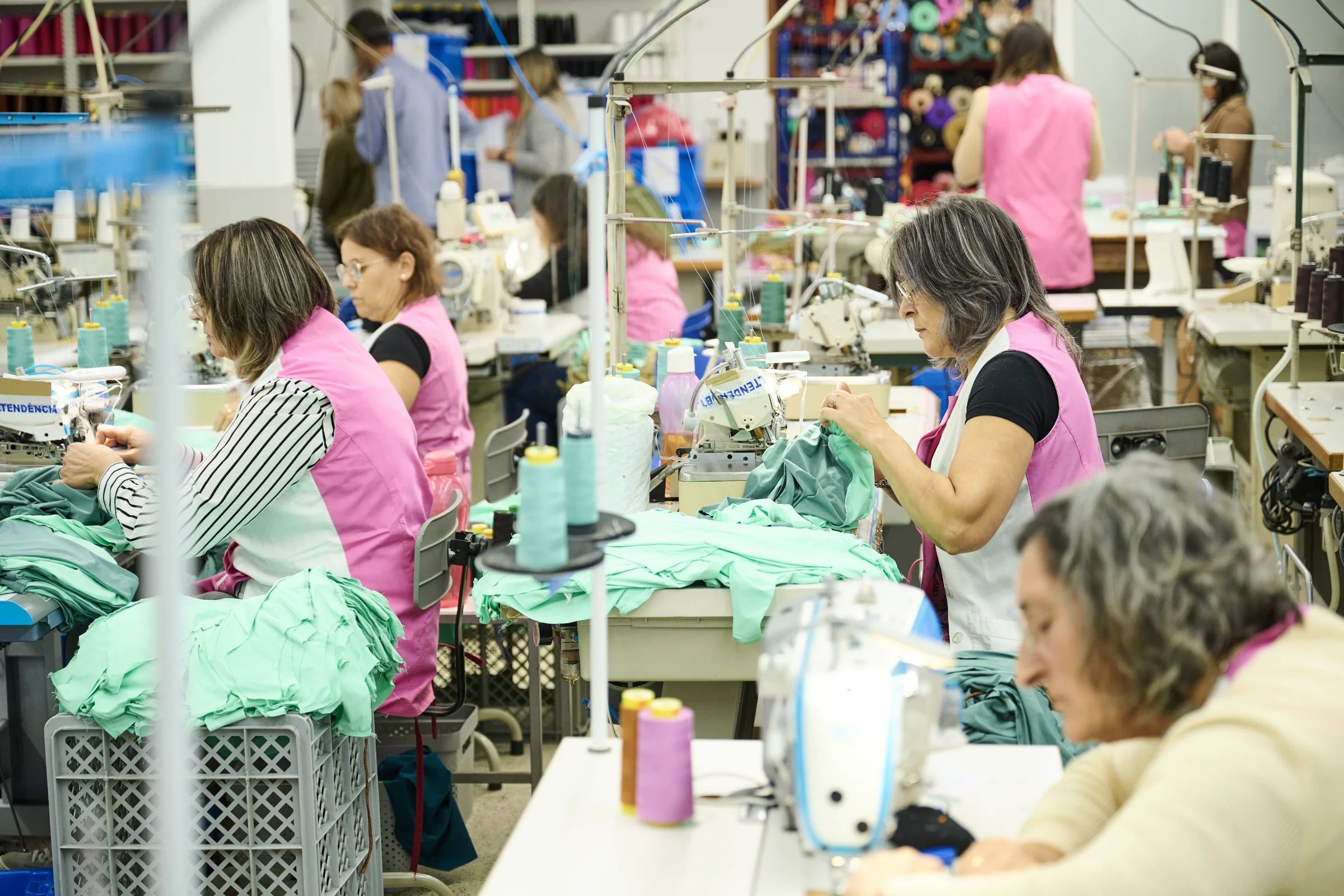 Mehrere Näherinnen in pinken Westen arbeiten in einer hellen Textilfabrik an Nähmaschinen, umgeben von türkisfarbenen Stoffen und Garnspulen.