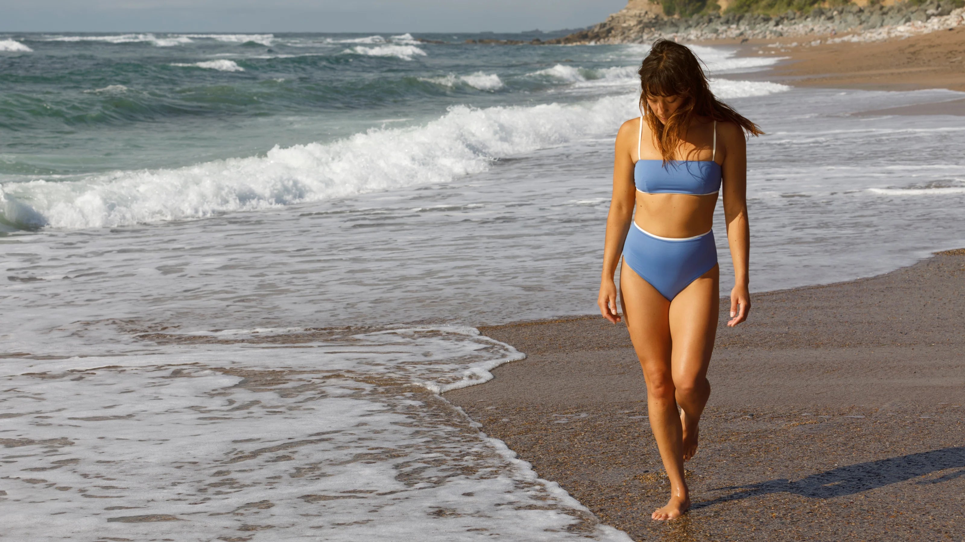 Frau im blauen Bikini spaziert barfuß am sandigen Meeresstrand.