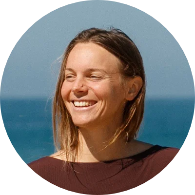 Lachende Frau mit offenen Haaren steht vor blauem Meer im Sonnenlicht.