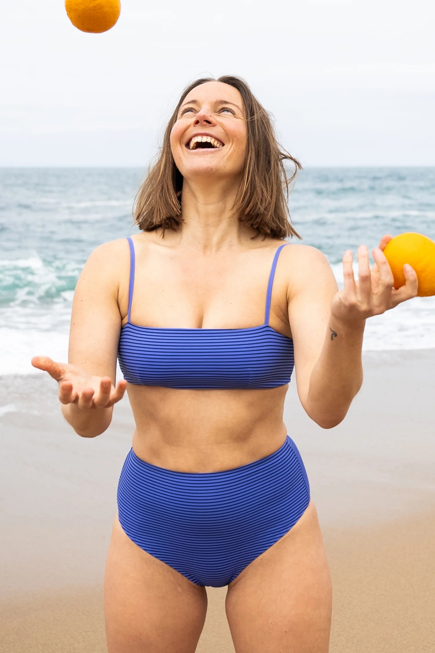 Frau am Strand in blauem MYMARINI-Bikini mit Spaghettiträgern und High-Waist-Schnitt jongliert mit Orangen.#color_blueberry-striped