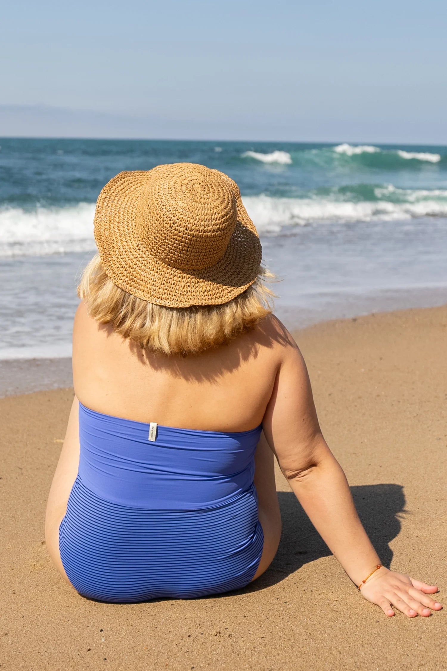 Person am Strand in einem blauen MYMARINI-Badeanzug mit Bandeau-Schnitt, sitzt im Sand und blickt aufs Meer.#color_blueberry-striped