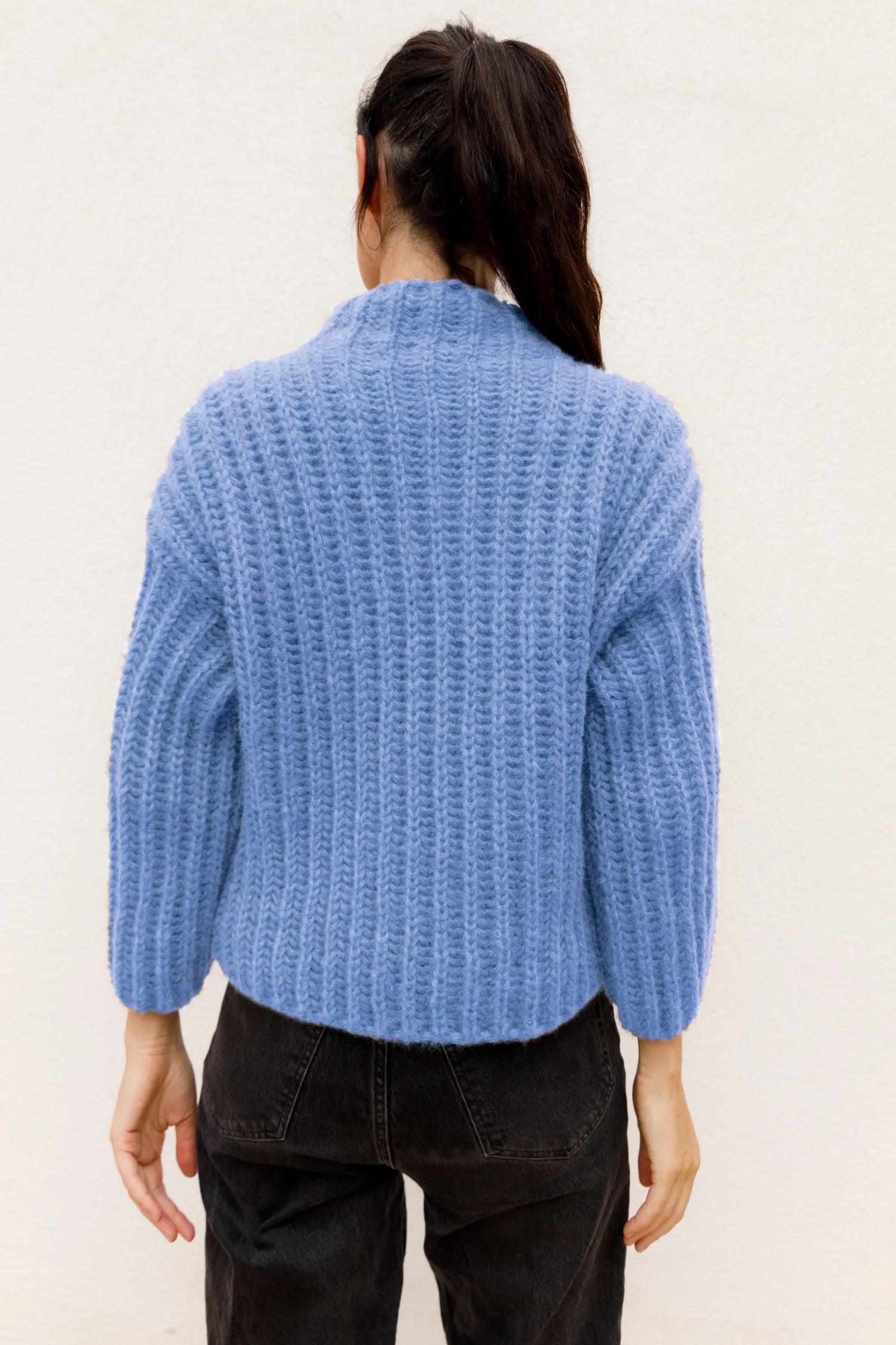 Frau in blauem Strickpullover und schwarzer Hose von hinten, Haare im Pferdeschwanz.#color_capri