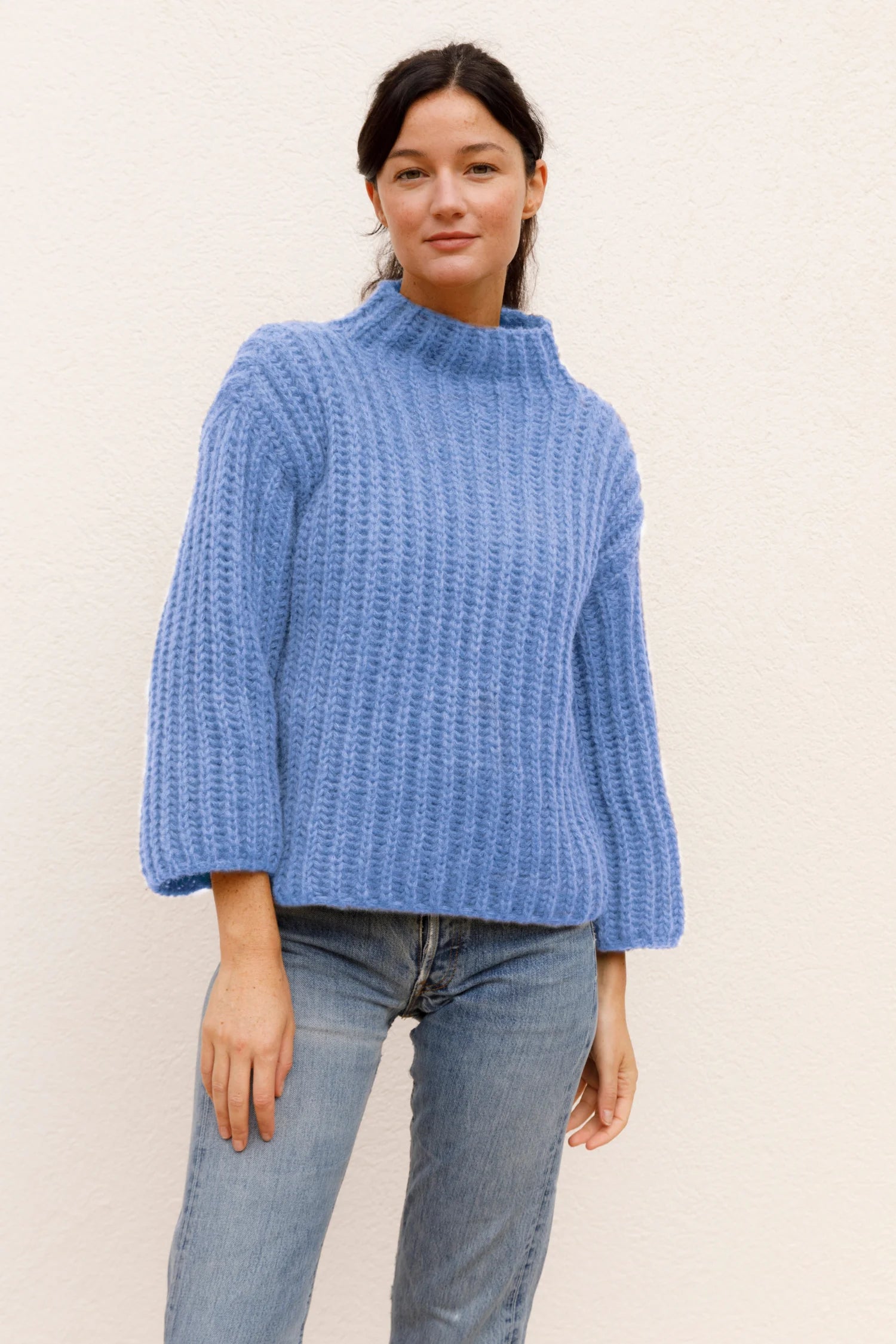 Frau in blauem Strickpullover und Jeans vor hellem Hintergrund.#color_capri