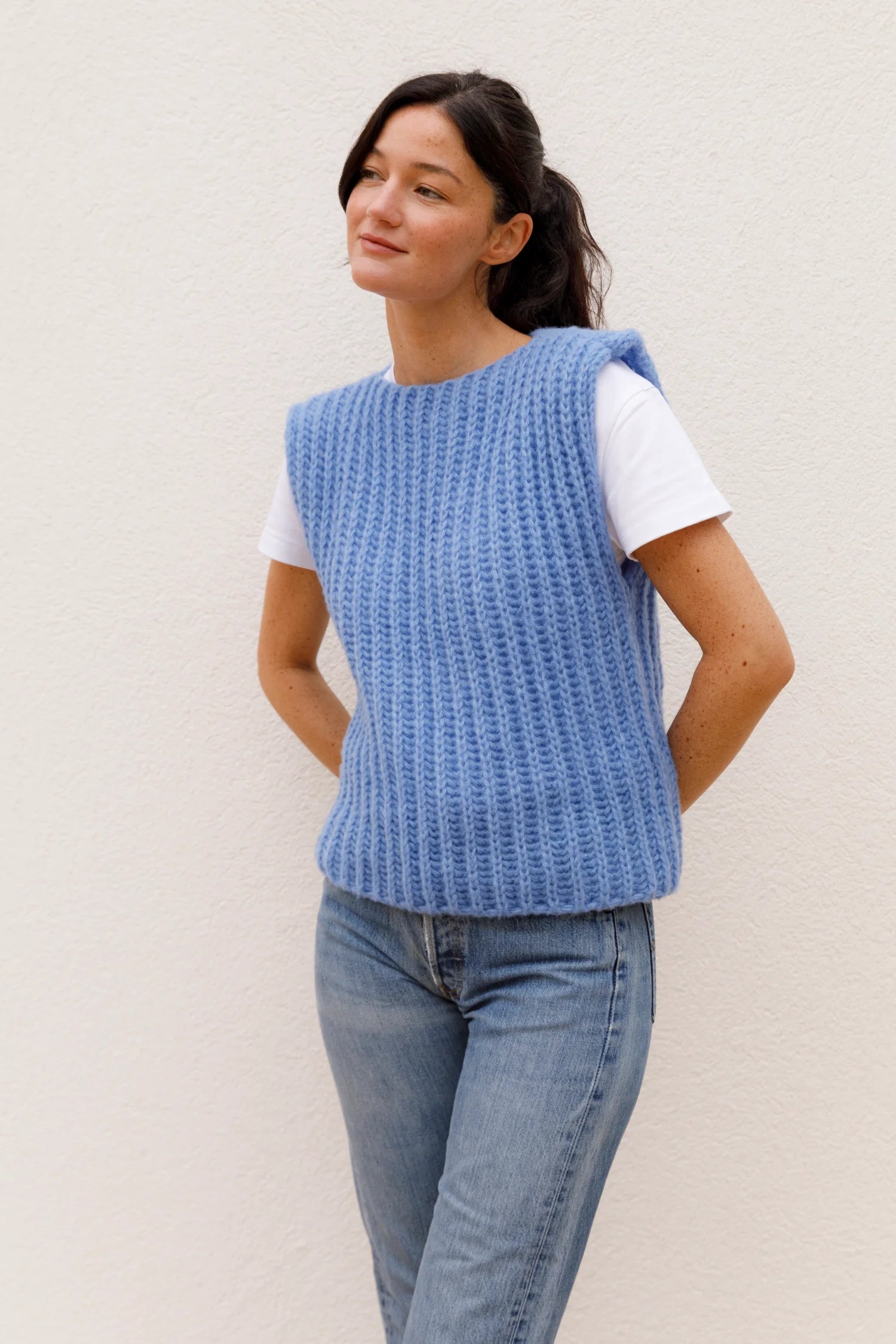 Frau in blauer Strickweste über weißem T-Shirt und Jeans vor hellem Hintergrund.#color_capri