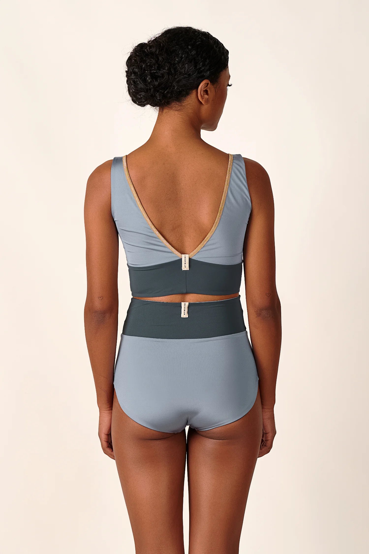 Rückansicht einer Frau in einem MYMARINI-Bikinitop mit V-Ausschnitt und einer High-Waist-Bikinihose in Blau.#color_crystal-grey