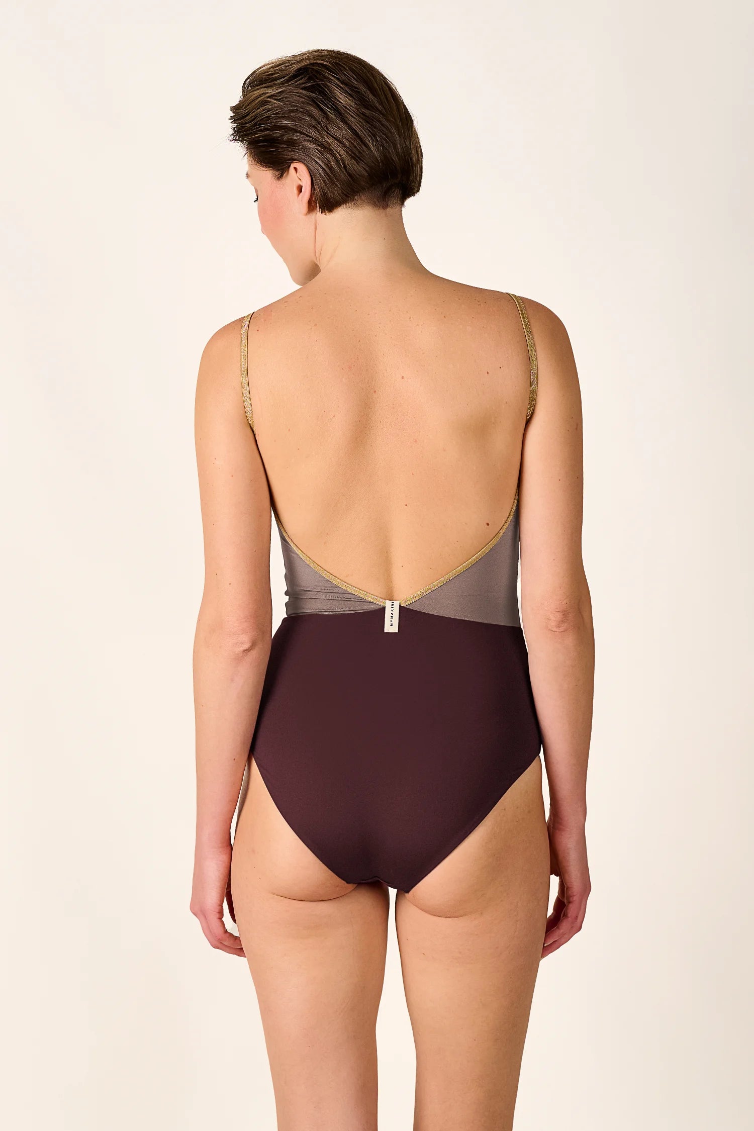Person von hinten in einem MYMARINI One-Piece mit tiefem Rückenausschnitt und Spaghettiträgern in Braun und Grau.#color_shimmer-brown