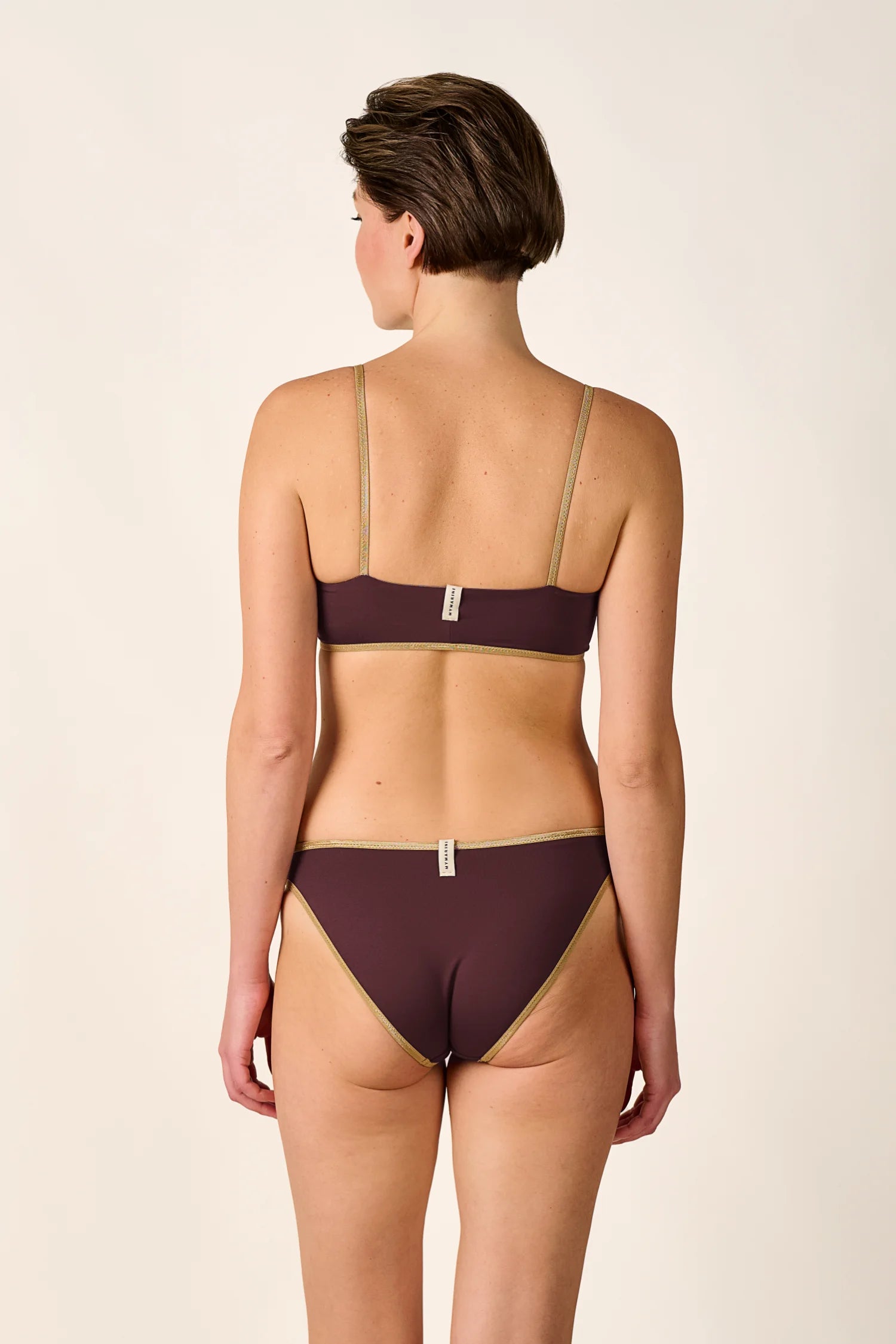 Frau in MYMARINI Bikini mit Spaghettiträgern und hohem Beinausschnitt, Rückansicht, vor neutralem Hintergrund.#color_shimmer-brown