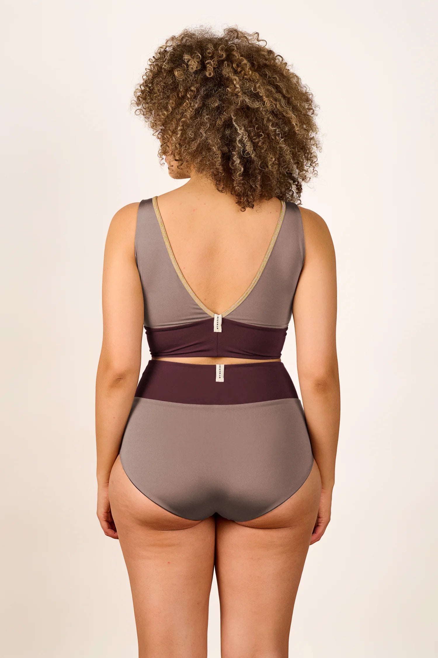 Person trägt einen MYMARINI Badeanzug mit V-Ausschnitt und hohem Beinausschnitt in Erdtönen, von hinten gezeigt.#color_shimmer-brown