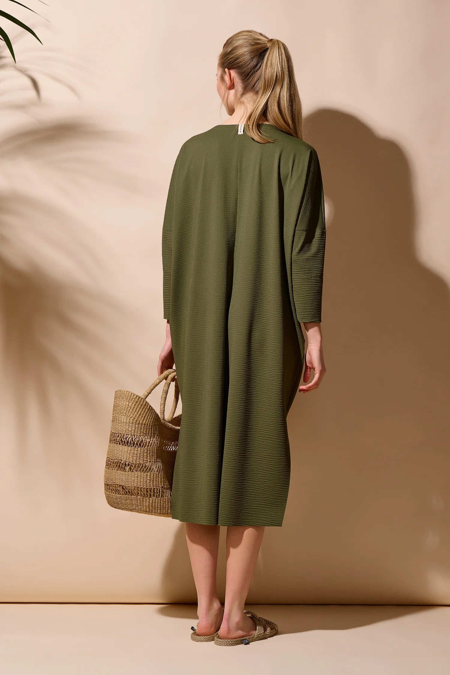 Person in langem, olivgrünem MYMARINI-Kleid mit langen Ärmeln, hält eine Strohtasche, steht vor beigem Hintergrund.#color_seaweed