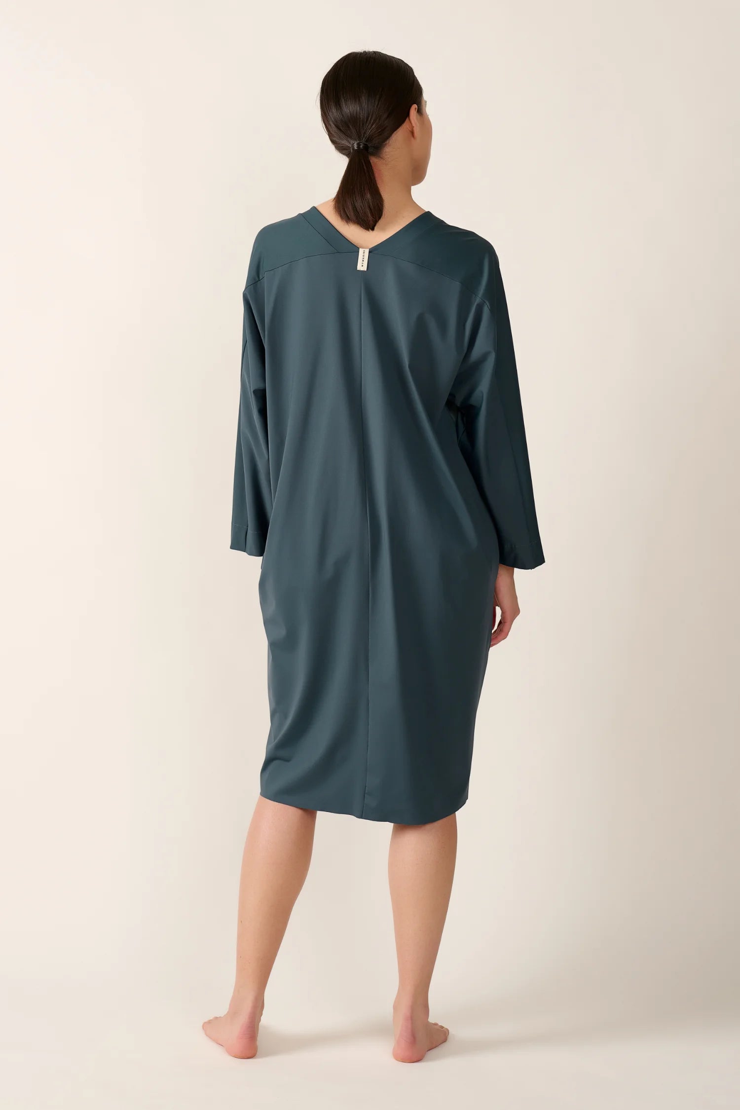 Person in lockerem, knielangem MYMARINI-Kleid mit V-Ausschnitt und langen Ärmeln, von hinten gezeigt.#color_grey