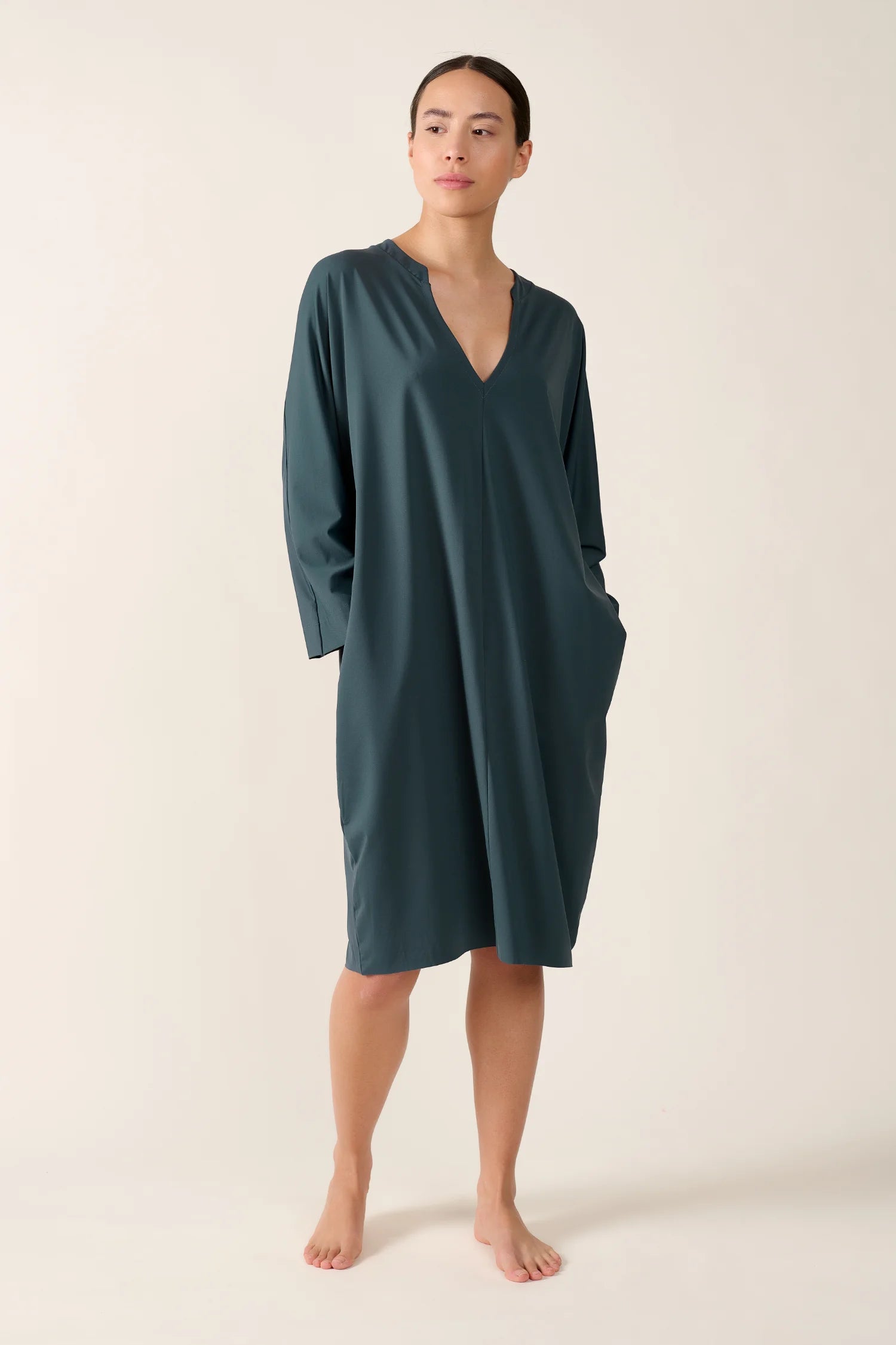 Person trägt ein MYMARINI-Kleid mit V-Ausschnitt in dunklem Grün, stehend vor neutralem Hintergrund.#color_grey