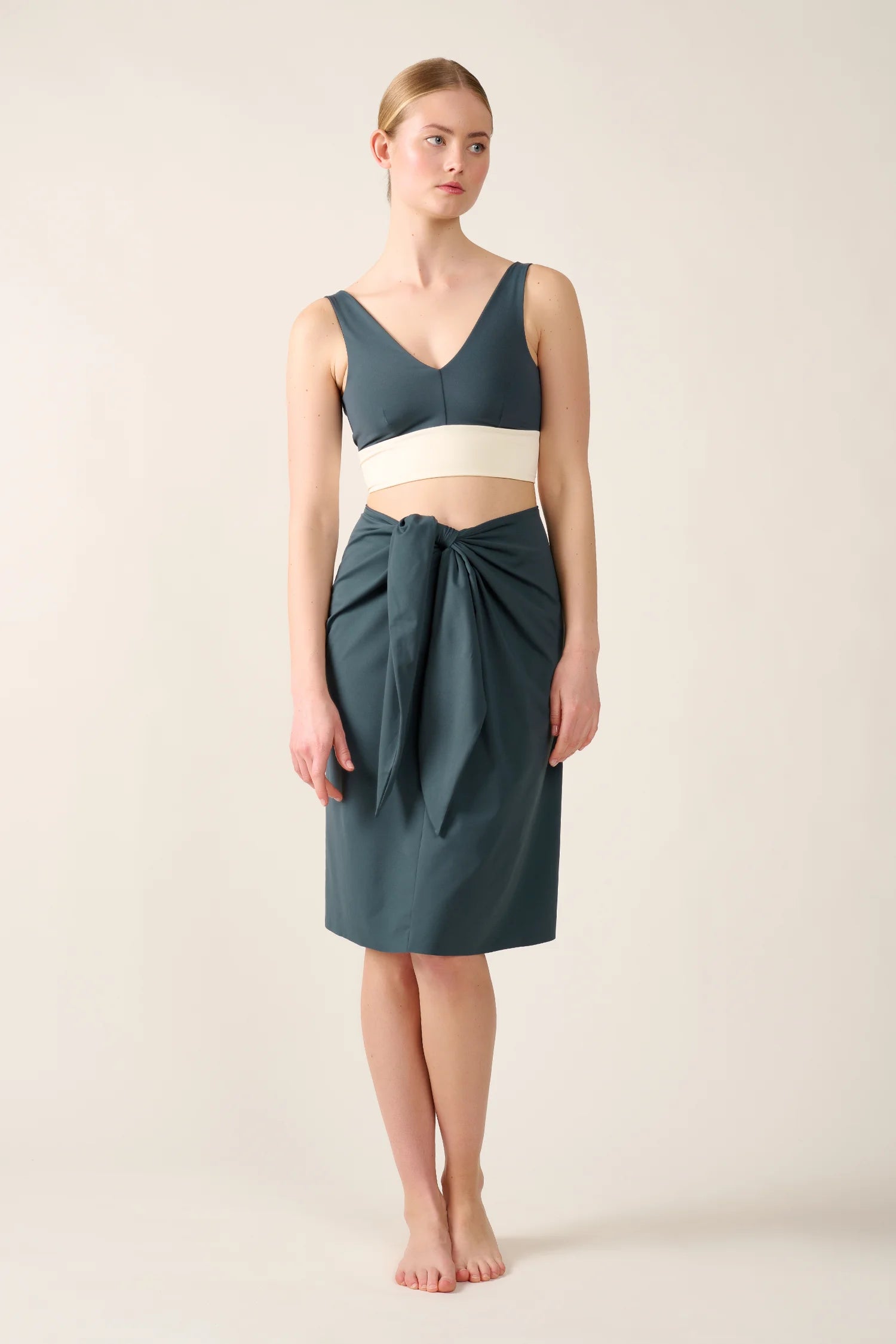 Frau trägt MYMARINI Bikinitop mit V-Ausschnitt und Mid-rise Rock in Dunkelgrün, stehend vor neutralem Hintergrund.#color_grey