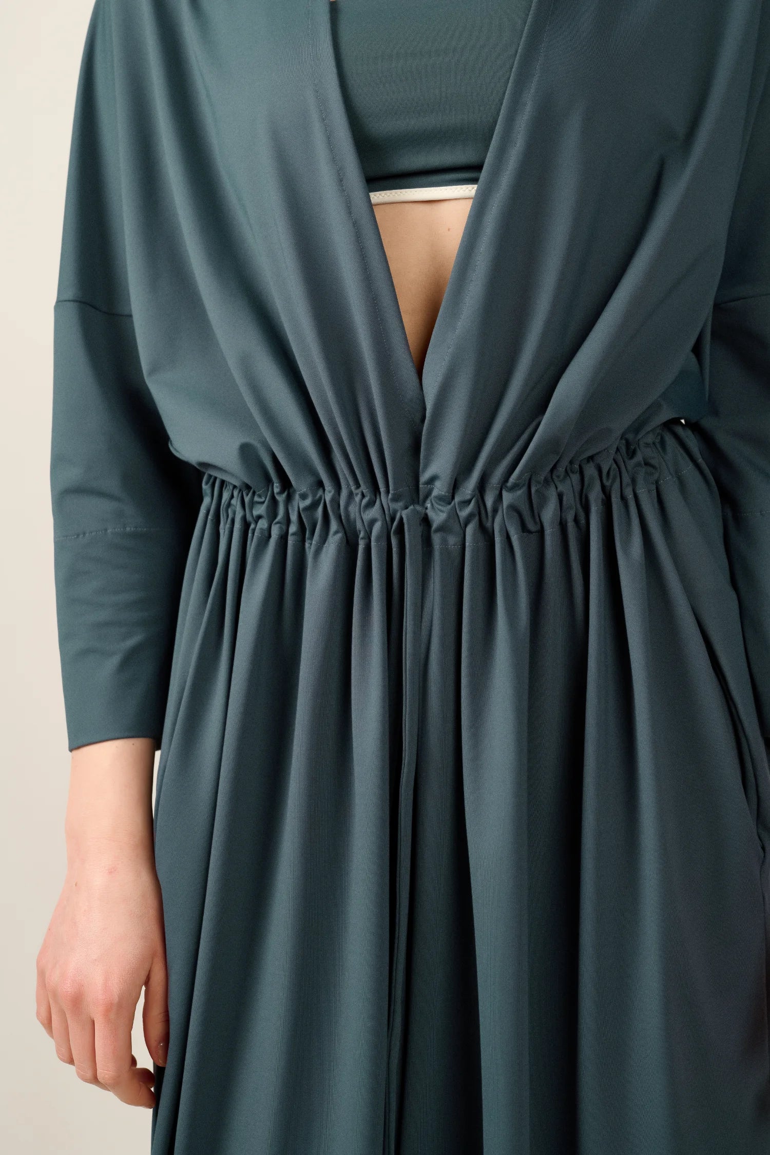 Person trägt ein MYMARINI-Kleid mit tiefem V-Ausschnitt und geraffter Taille. Darunter ein hochgeschlossenes Top sichtbar.#color_grey