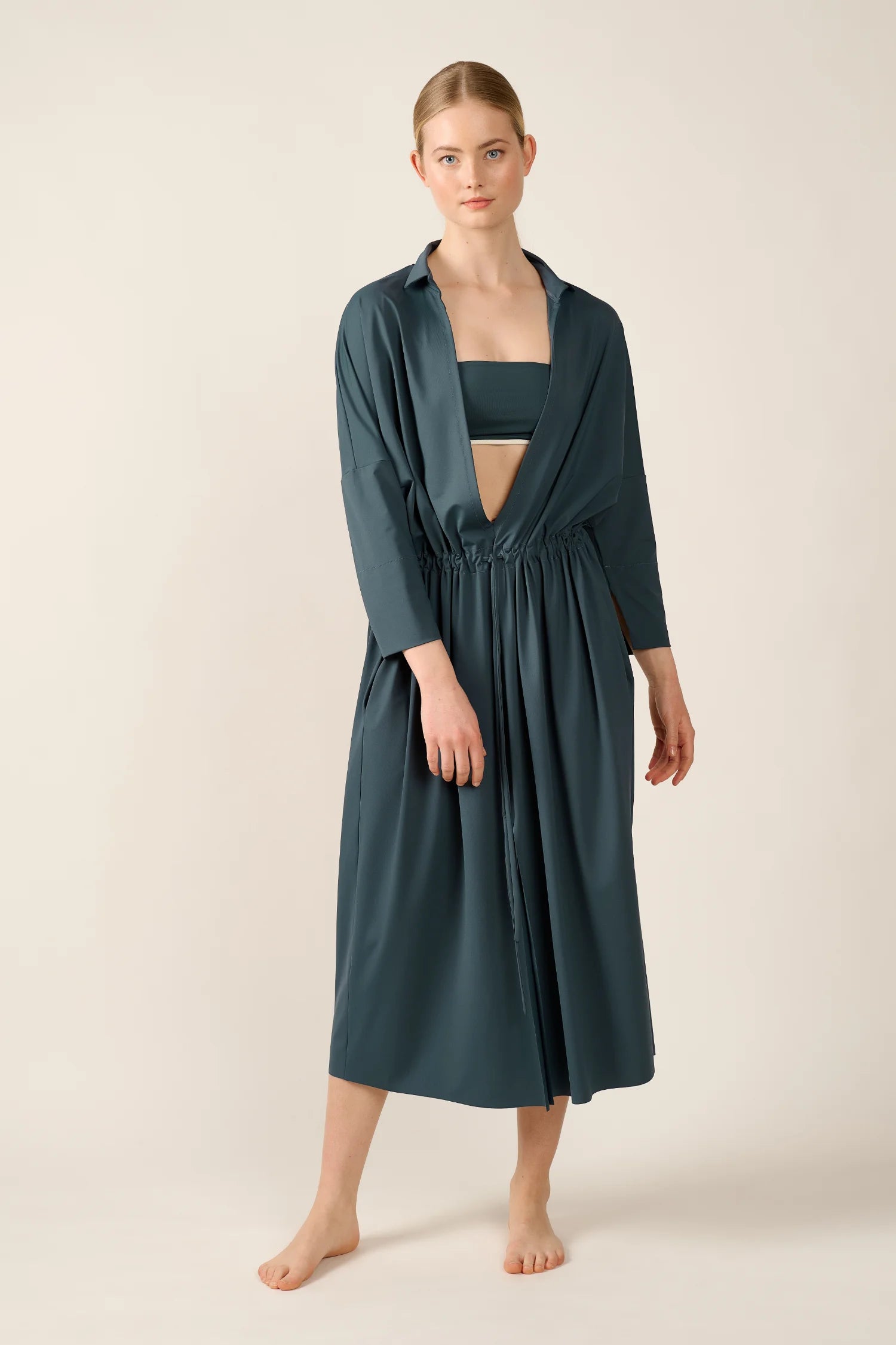 Frau trägt MYMARINI Badeanzug mit Bandeau-Top und tiefem V-Ausschnitt, kombiniert mit langem, lockerem Kleid.#color_grey