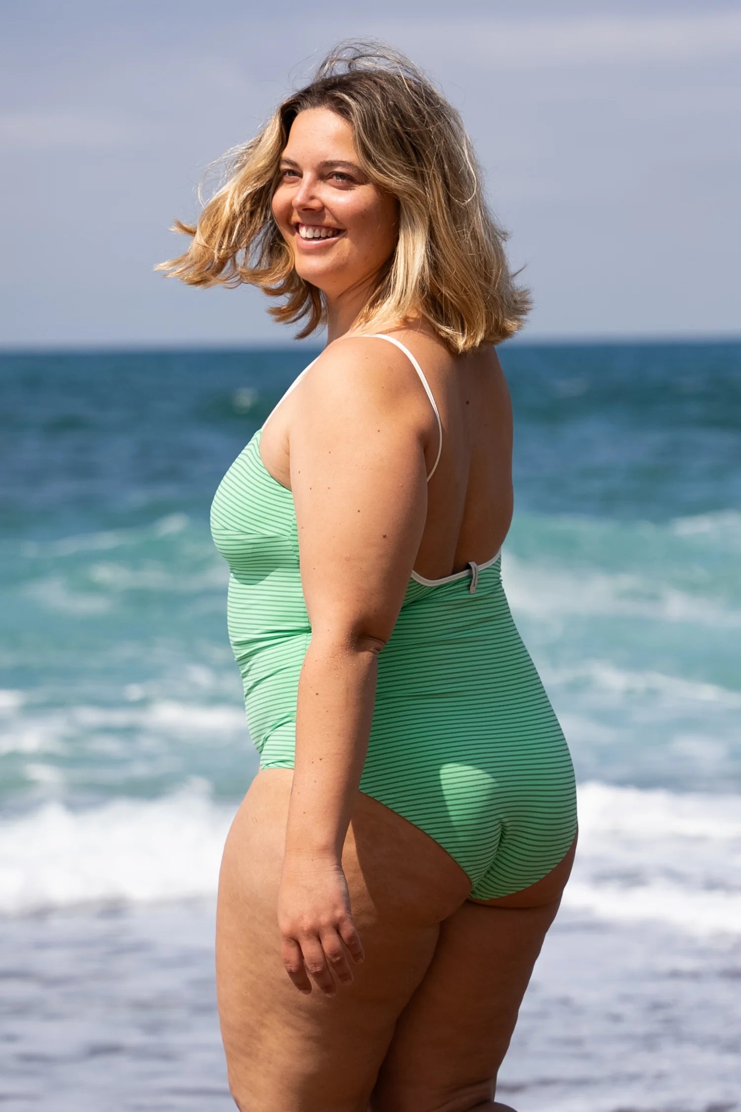 Frau am Strand in einem MYMARINI Badeanzug mit tiefem Rückenausschnitt und breiten Trägern, lächelnd vor dem Meer.#color_tonic-striped
