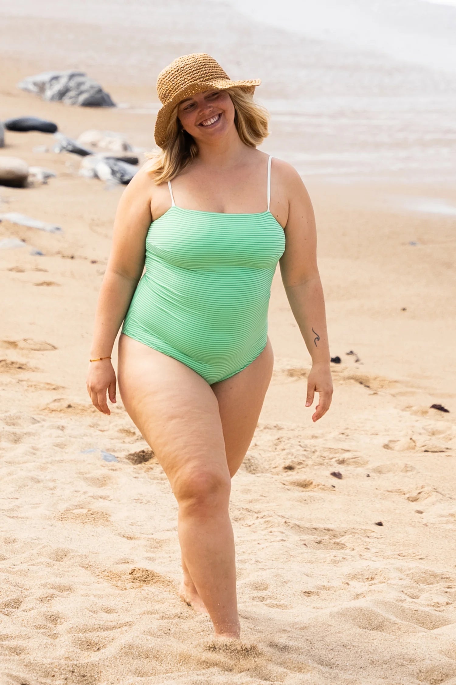 Frau am Strand in einem MYMARINI Badeanzug mit Spaghettiträgern, lächelnd und einen Strohhut tragend.#color_tonic-striped