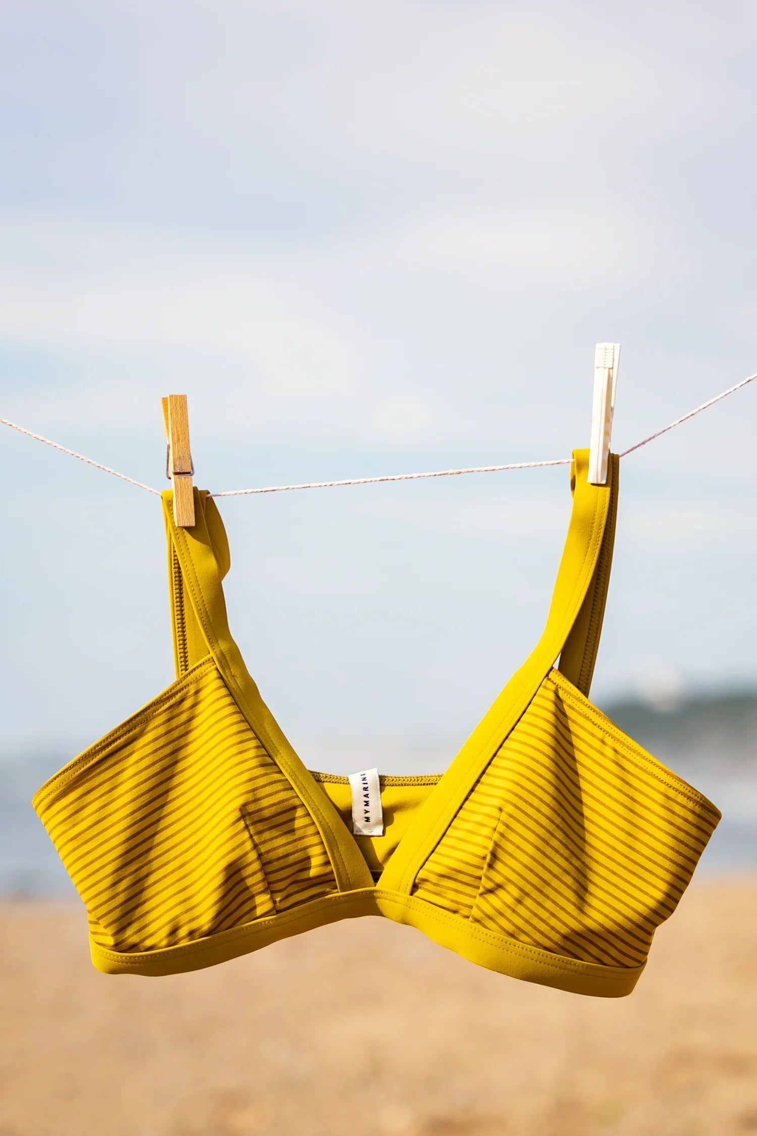 Gelbes Bikinitop mit schwarzem Muster von MYMARINI in Vorderansicht, aufgehängt an einer Leine am Strand.#color_pear-striped
