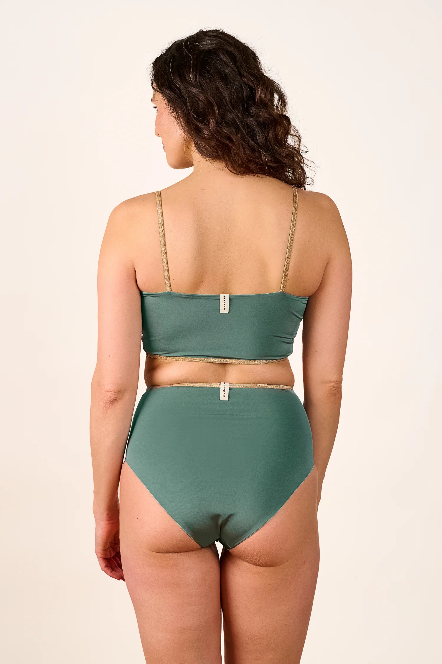 Frau trägt MYMARINI Bikini mit Bandeau-Top und High-Waist-Hose in Grün, Rückansicht, vor neutralem Hintergrund.#color_lagoon-teal
