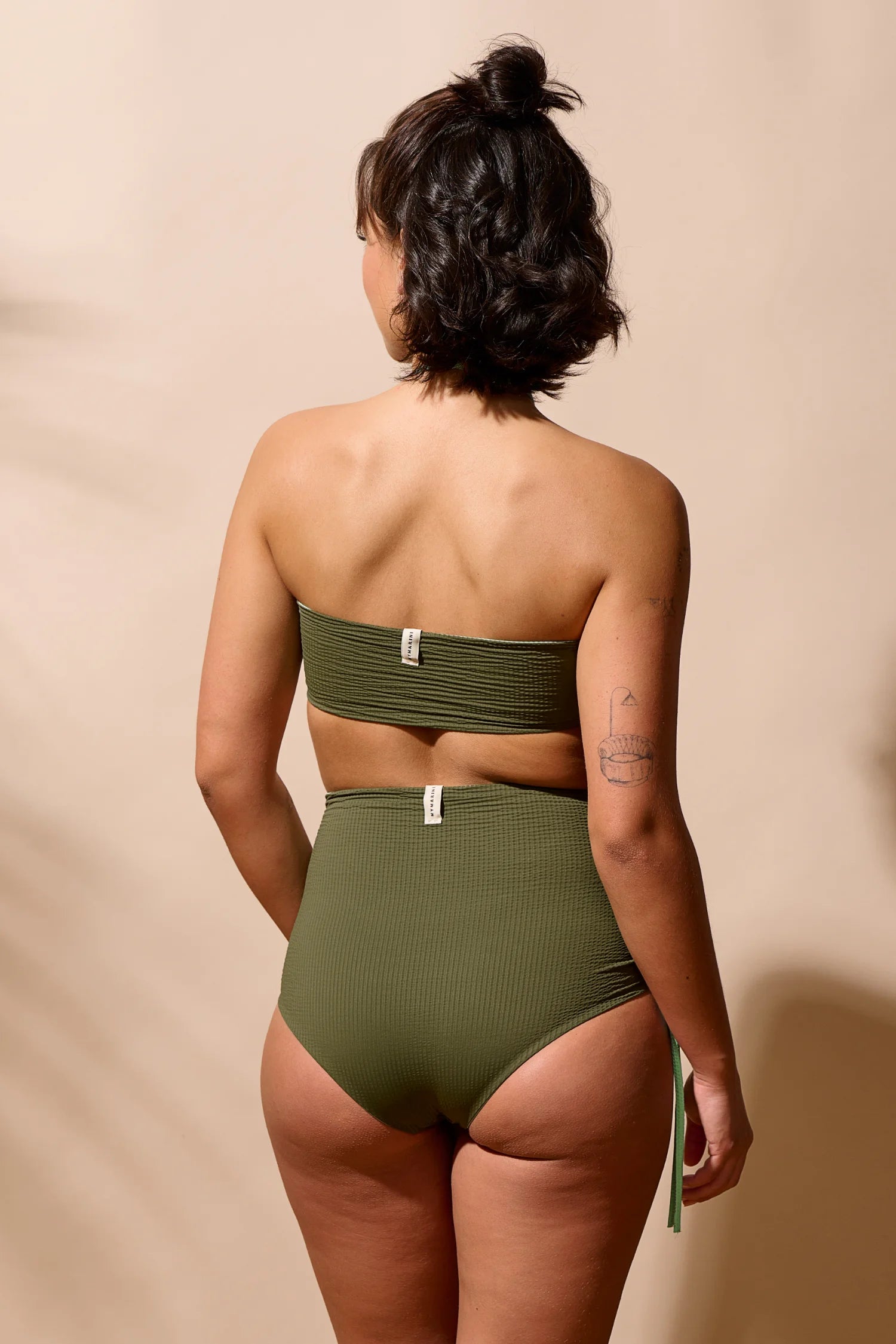 Frau in MYMARINI Bikini mit Bandeau-Top und High-Waist-Hose in Olivgrün, von hinten vor beigem Hintergrund.#color_lagoon-seaweed