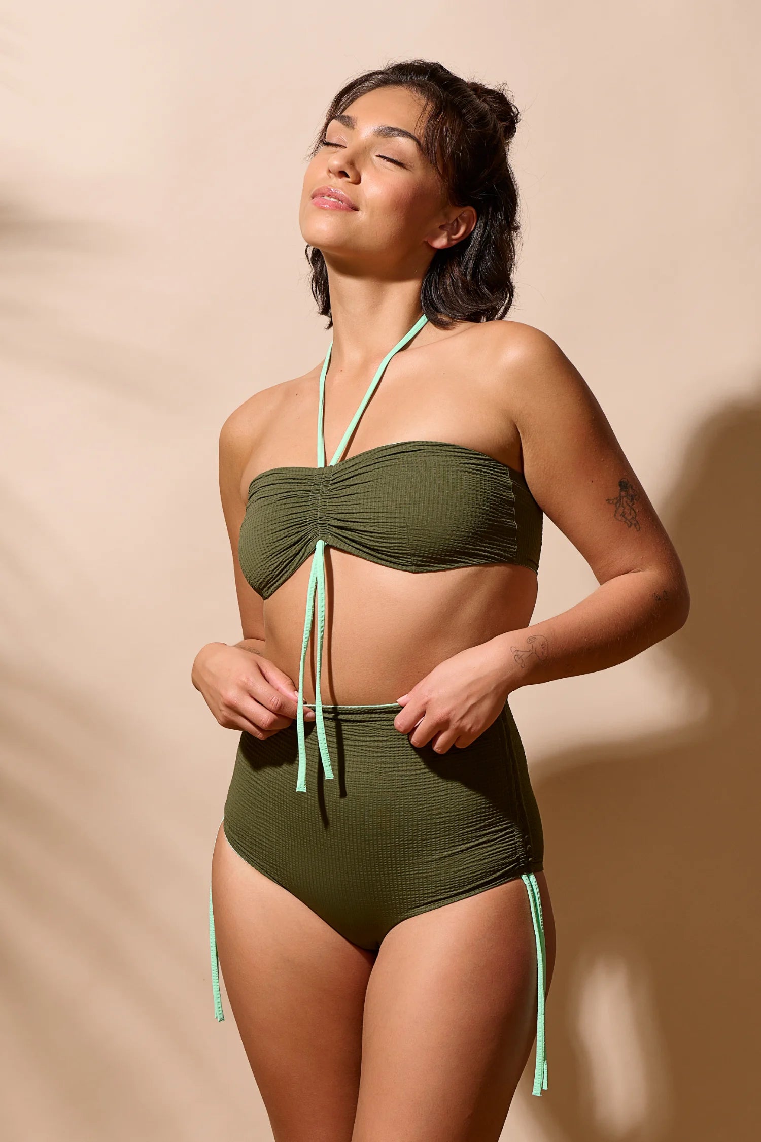 Frau trägt MYMARINI Bikini mit Bandeau-Top und High-Waist-Hose in Olivgrün, vor neutralem Hintergrund.#color_lagoon-seaweed