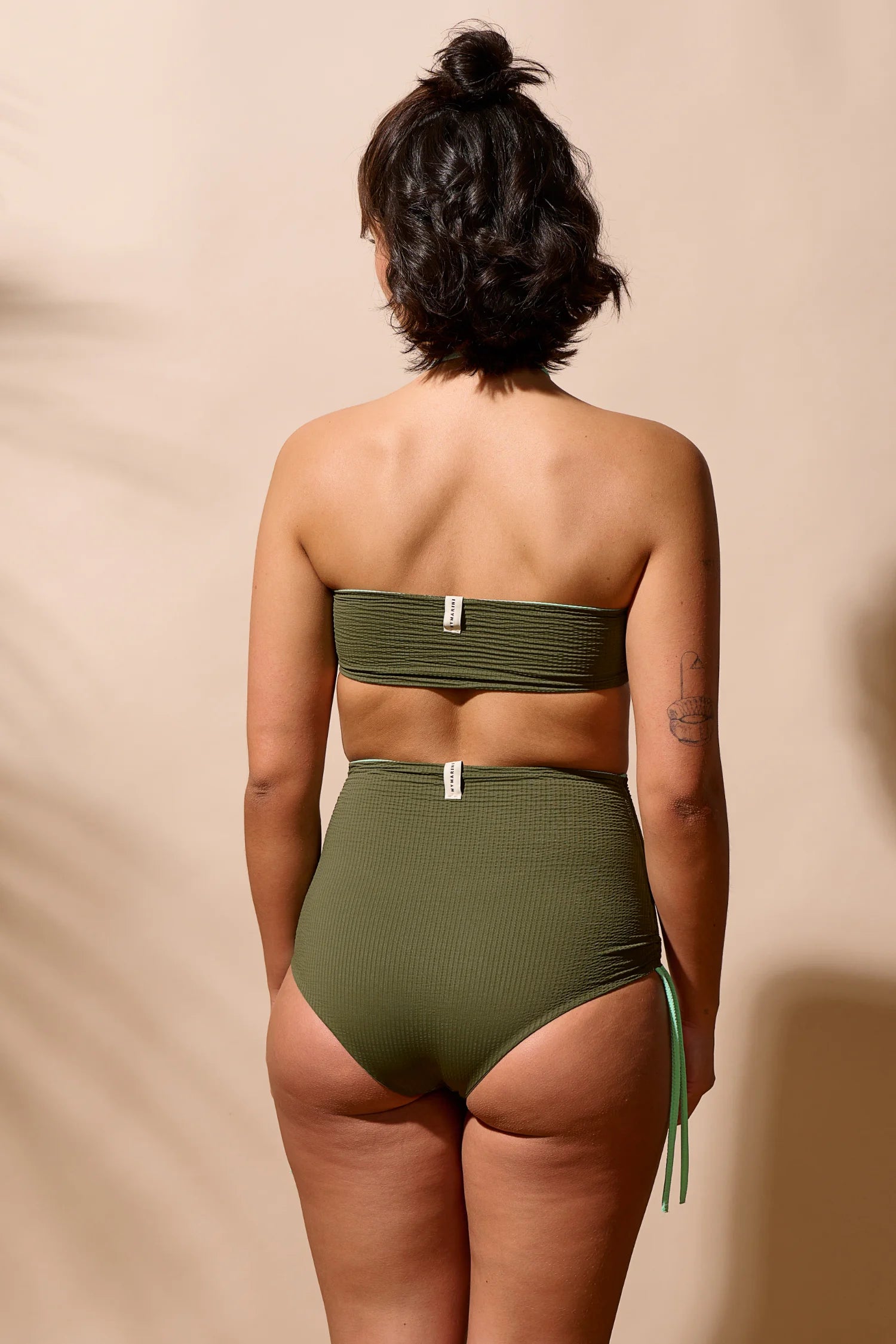 Frau trägt MYMARINI Bikini mit Bandeau-Top und High-Waist-Hose in Olivgrün, Rückansicht, vor beigem Hintergrund.#color_lagoon-seaweed