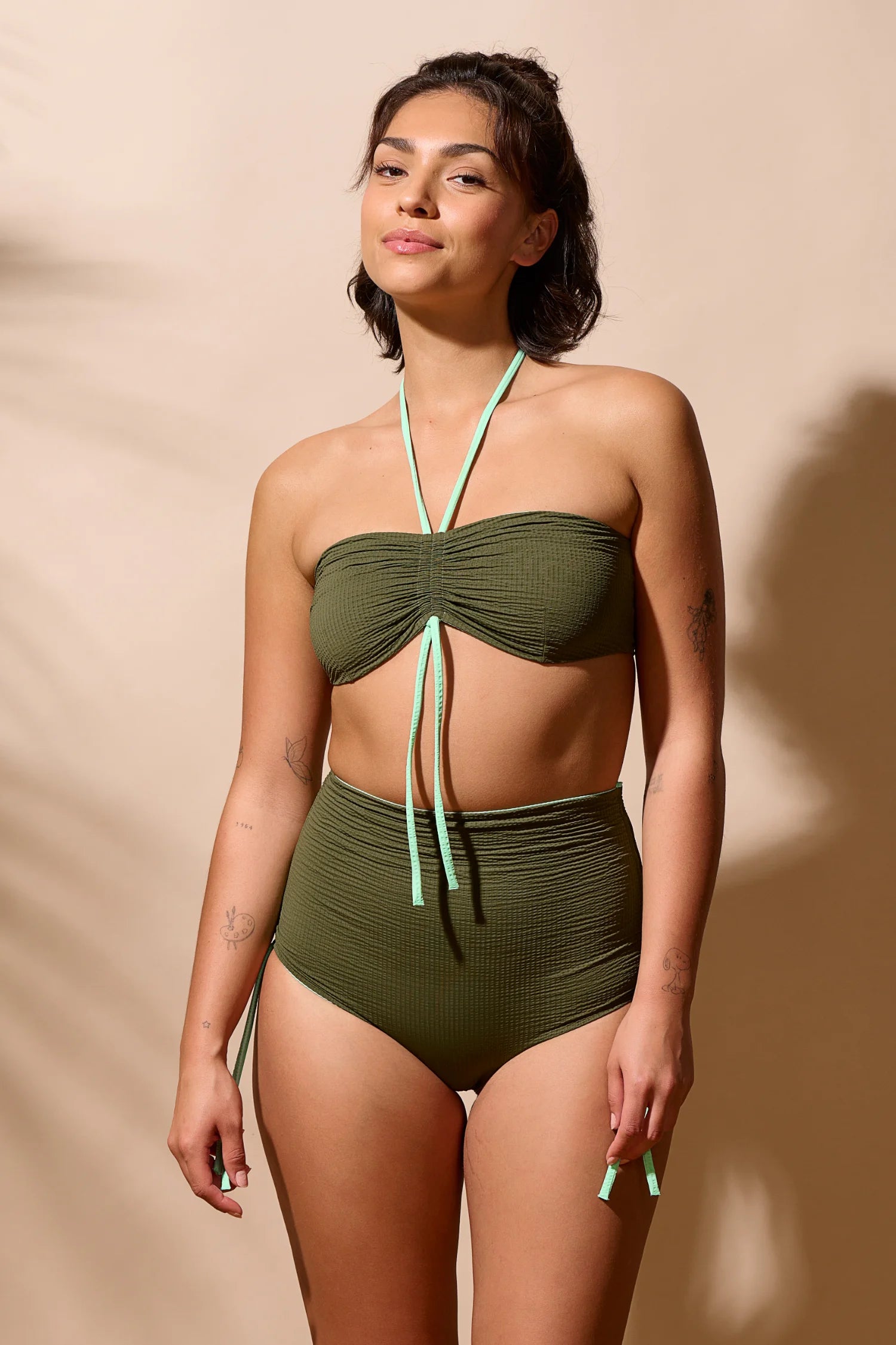 Frau trägt MYMARINI Bikini mit Bandeau-Top und High-Waist-Hose in Olivgrün vor beigem Hintergrund.#color_lagoon-seaweed