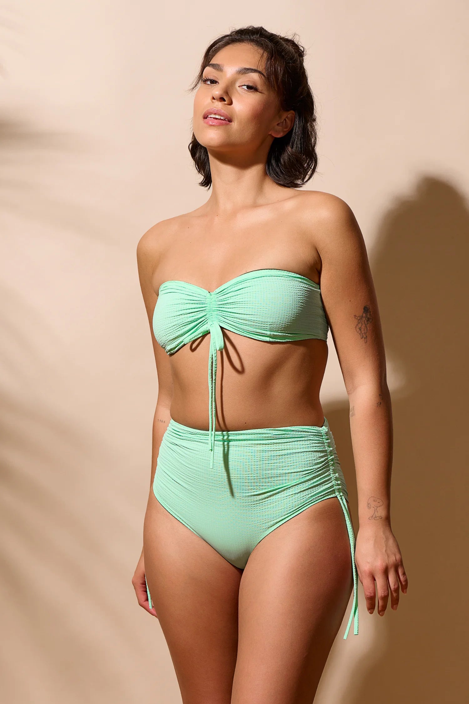Frau trägt MYMARINI Bikini mit Bandeau-Top und High-Waist-Hose in Pastellgrün vor beigem Hintergrund.#color_lagoon-seaweed