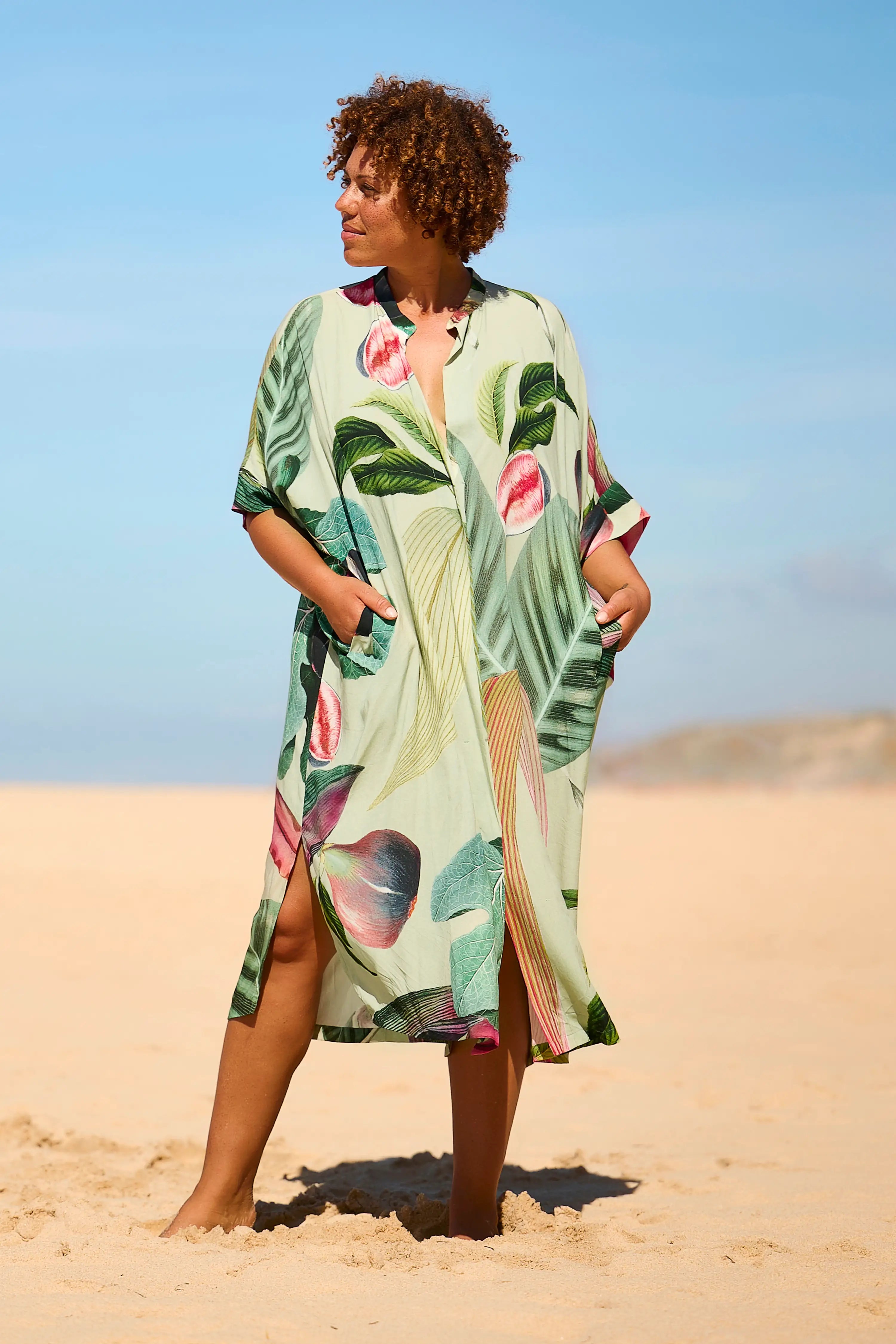 Frau am Strand trägt ein MYMARINI-Kleid mit tropischem Muster und kurzen Ärmeln.#color_tropical-print