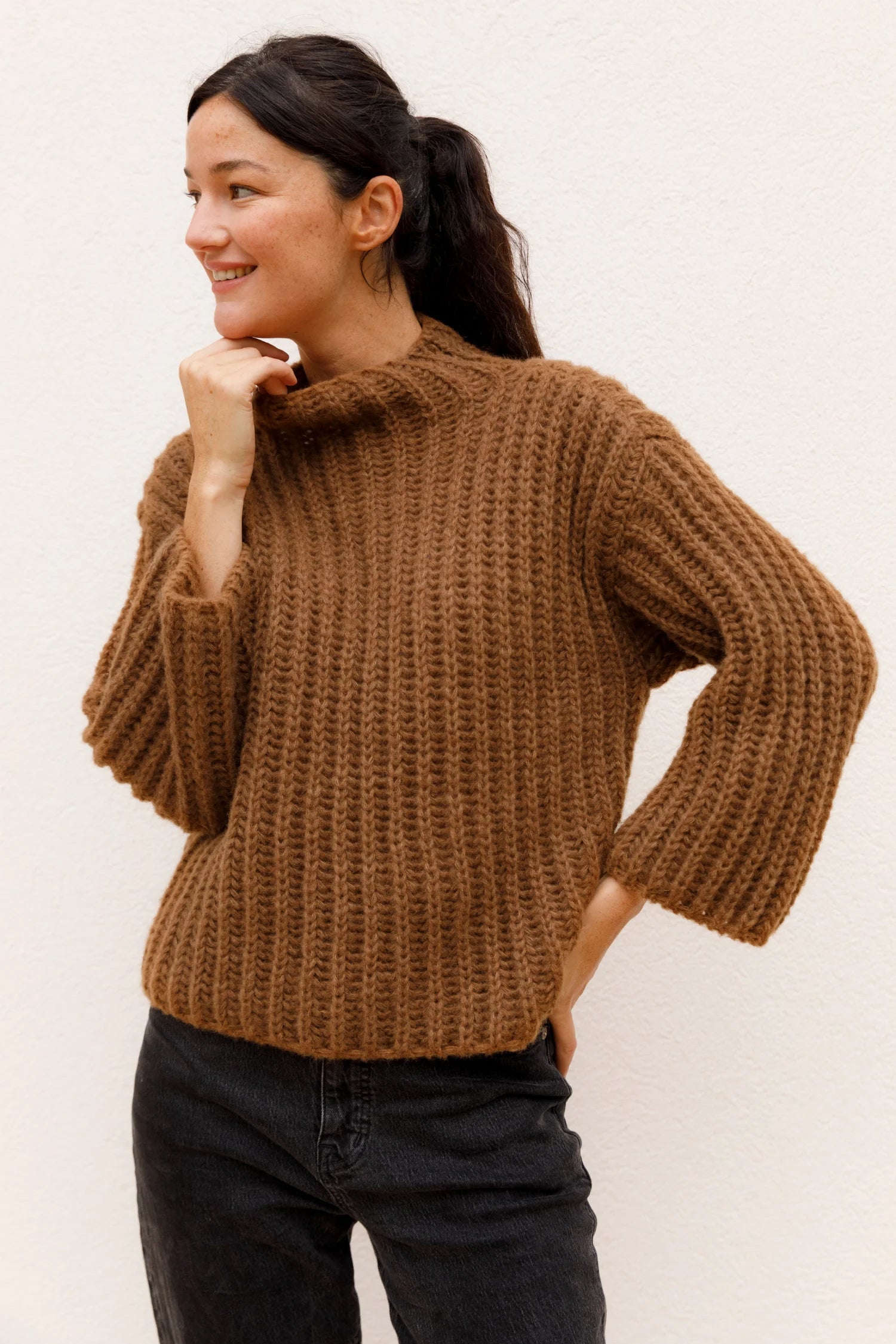 Person trägt einen braunen Strickpullover mit Stehkragen und schwarze Hose. Sie lächelt und blickt zur Seite.#color_camel