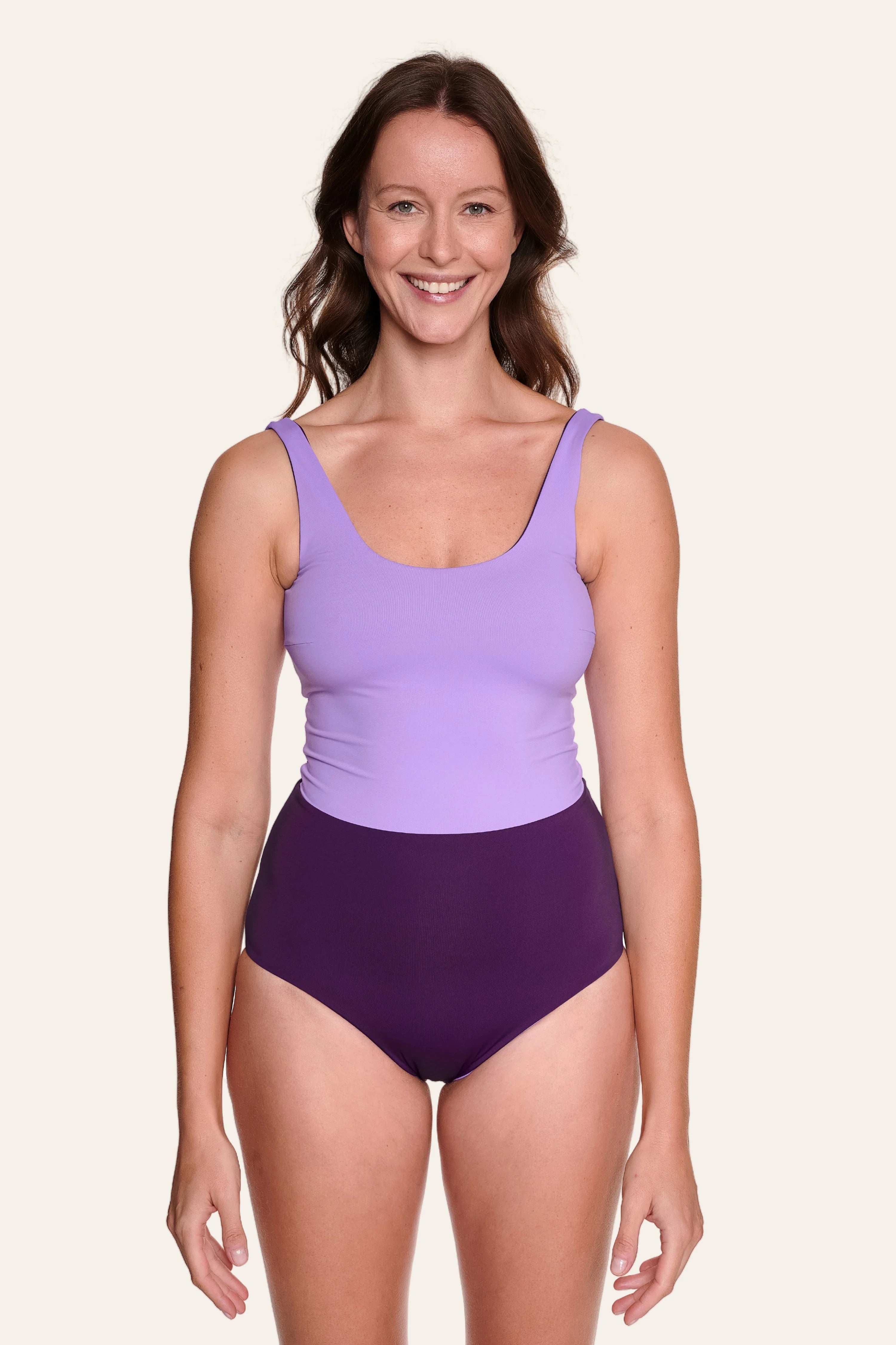 Frau trägt MYMARINI Badeanzug in Lila mit breiten Trägern und hohem Beinausschnitt, lächelnd vor hellem Hintergrund.#color_lavender-plum