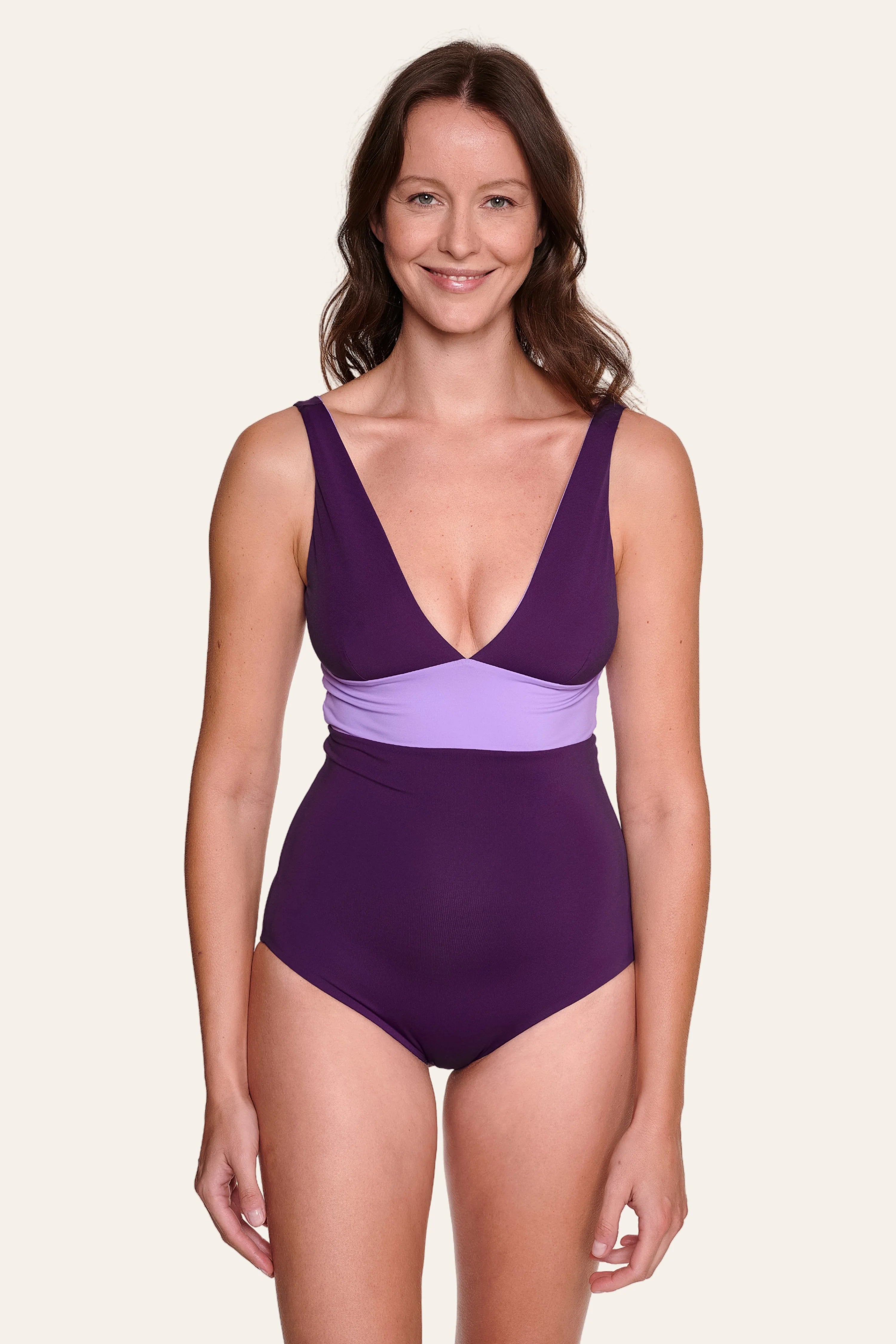 Frau trägt lila MYMARINI Badeanzug mit V-Ausschnitt und breiten Trägern, lächelnd vor hellem Hintergrund.#color_lavender-plum