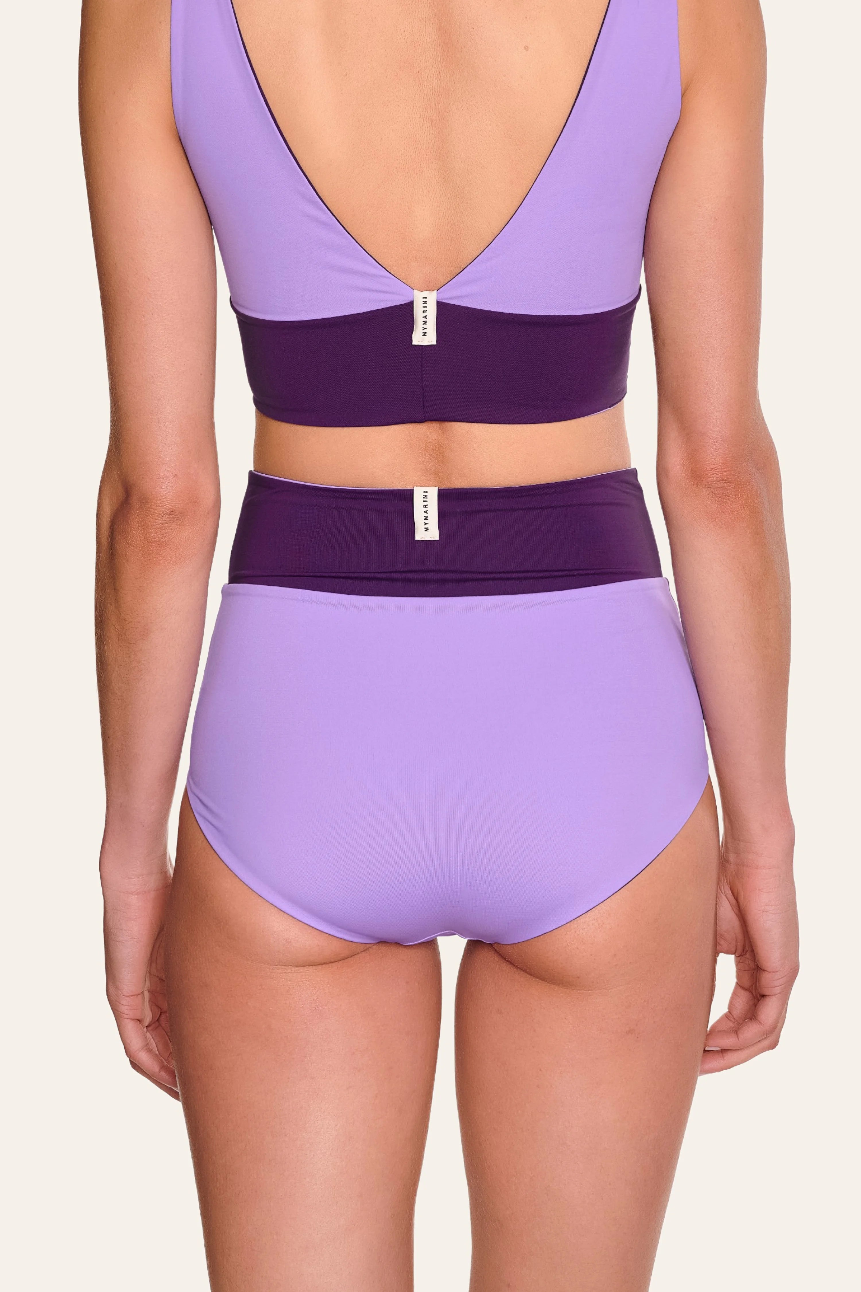 Frau trägt MYMARINI Bikini mit V-Ausschnitt und High Waist in Lila und Dunkelviolett, Rückansicht.#color_lavender-plum