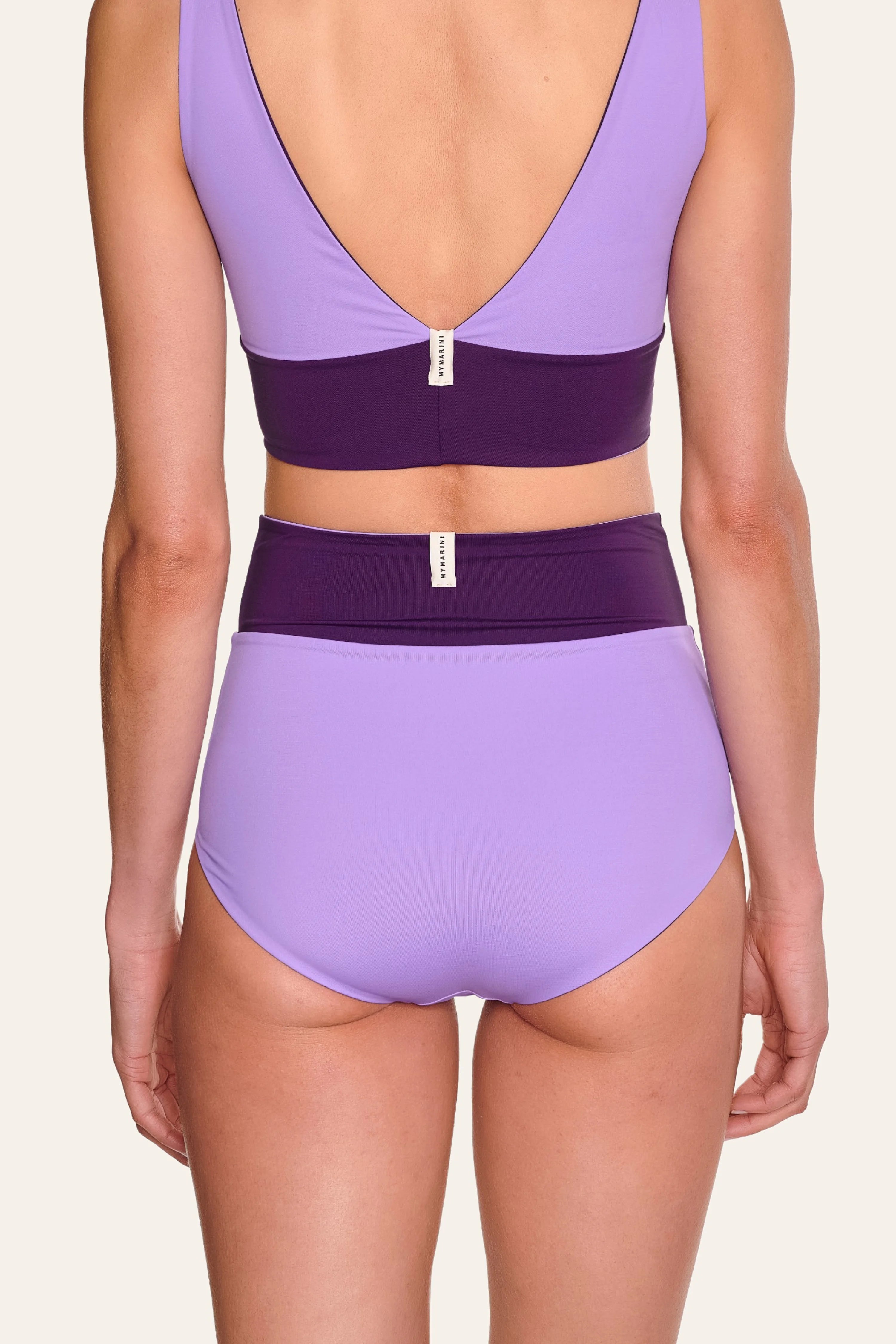 Rückansicht einer Person in MYMARINI Bikinihose mit hohem Bund und Bikinitop mit V-Ausschnitt in Lila und Dunkelviolett.#color_lavender-plum