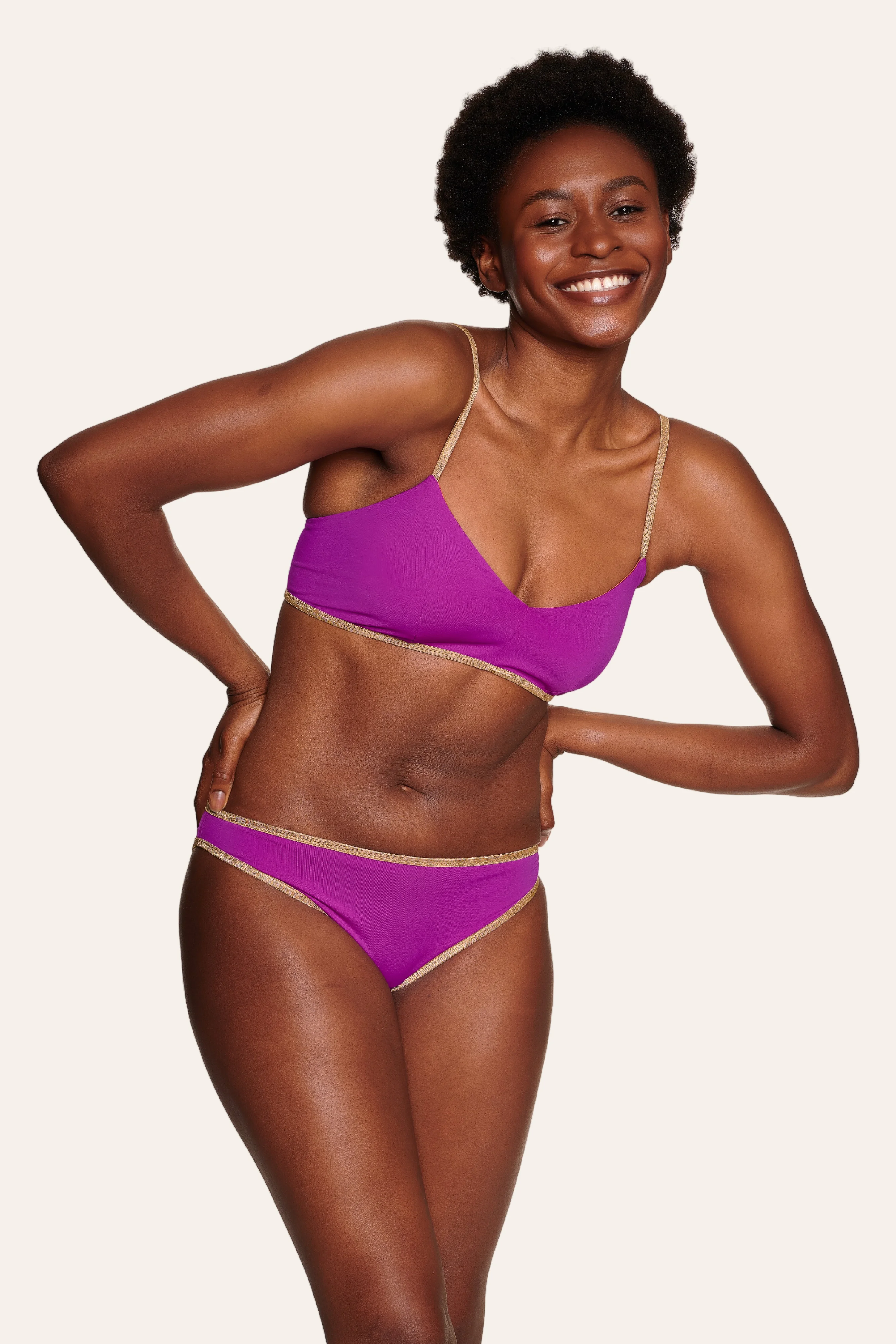 Frau in MYMARINI Bikini mit Spaghettiträgern und Mid rise Schnitt in Lila, lächelnd vor hellem Hintergrund.#color_papaya-cassis