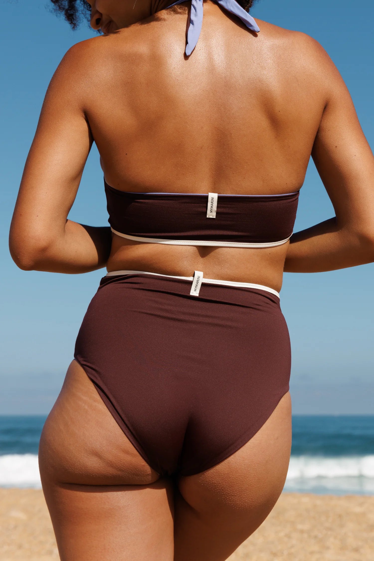 Rückseite eines braunen MYMARINI Bikini-Sets (Bandeau-Top mit Halsband, High-Waist-Unterteil) am Strand.#color_chocolate-lilac