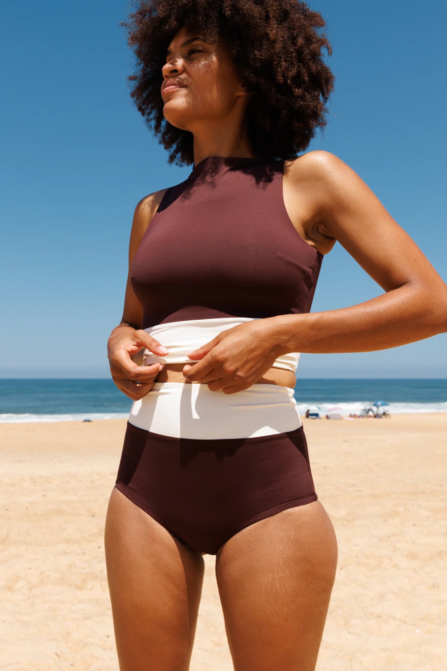 MYMARINI Bikini: High-Neck-Oberteil in Braun und High-Waist-Hose mit weißem Bund am Strand.#color_chocolate-lilac
