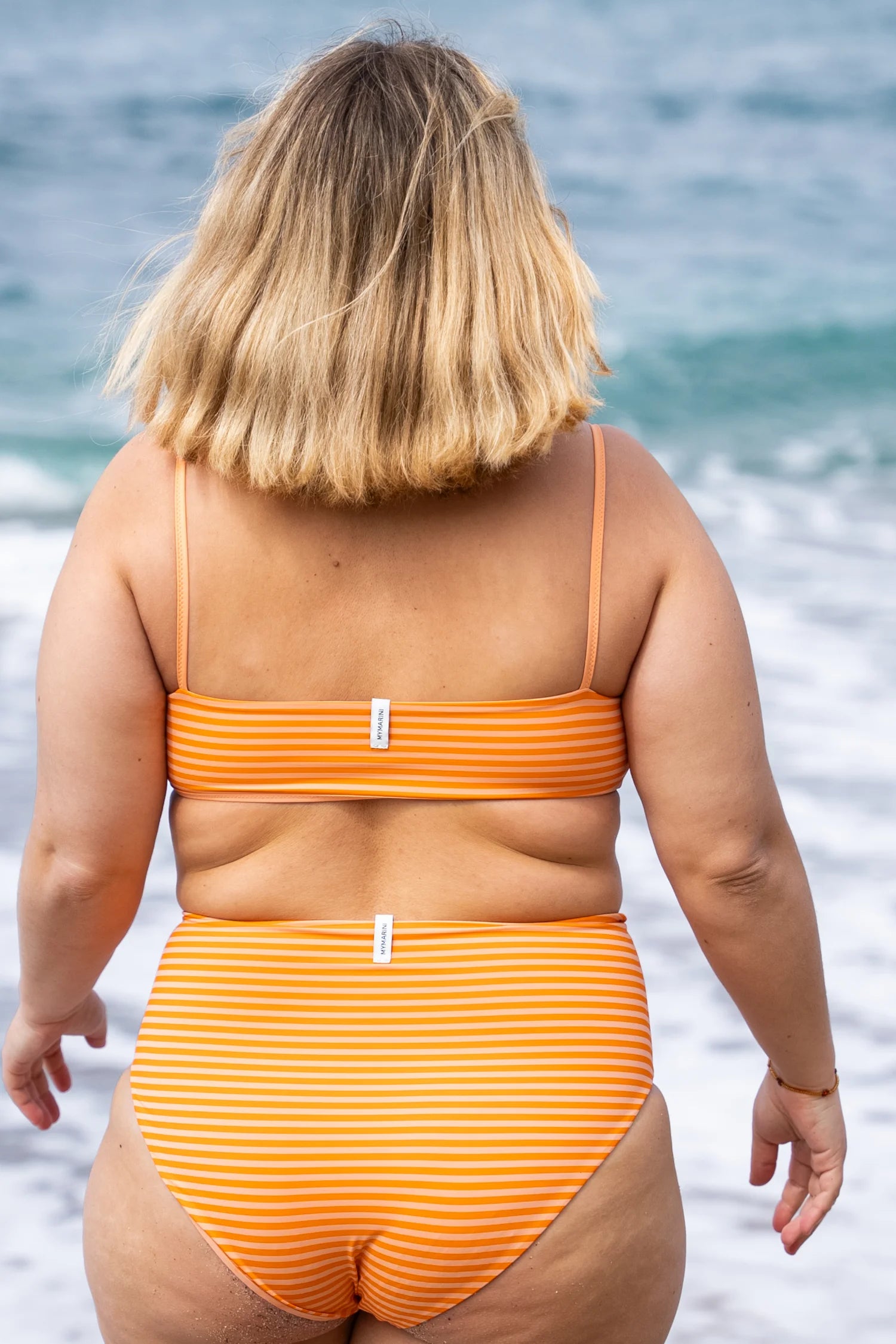 Person am Strand in gestreiftem MYMARINI Bikini mit Spaghettiträgern und High-Waist-Schnitt, Blick aufs Meer.#color_melon-striped