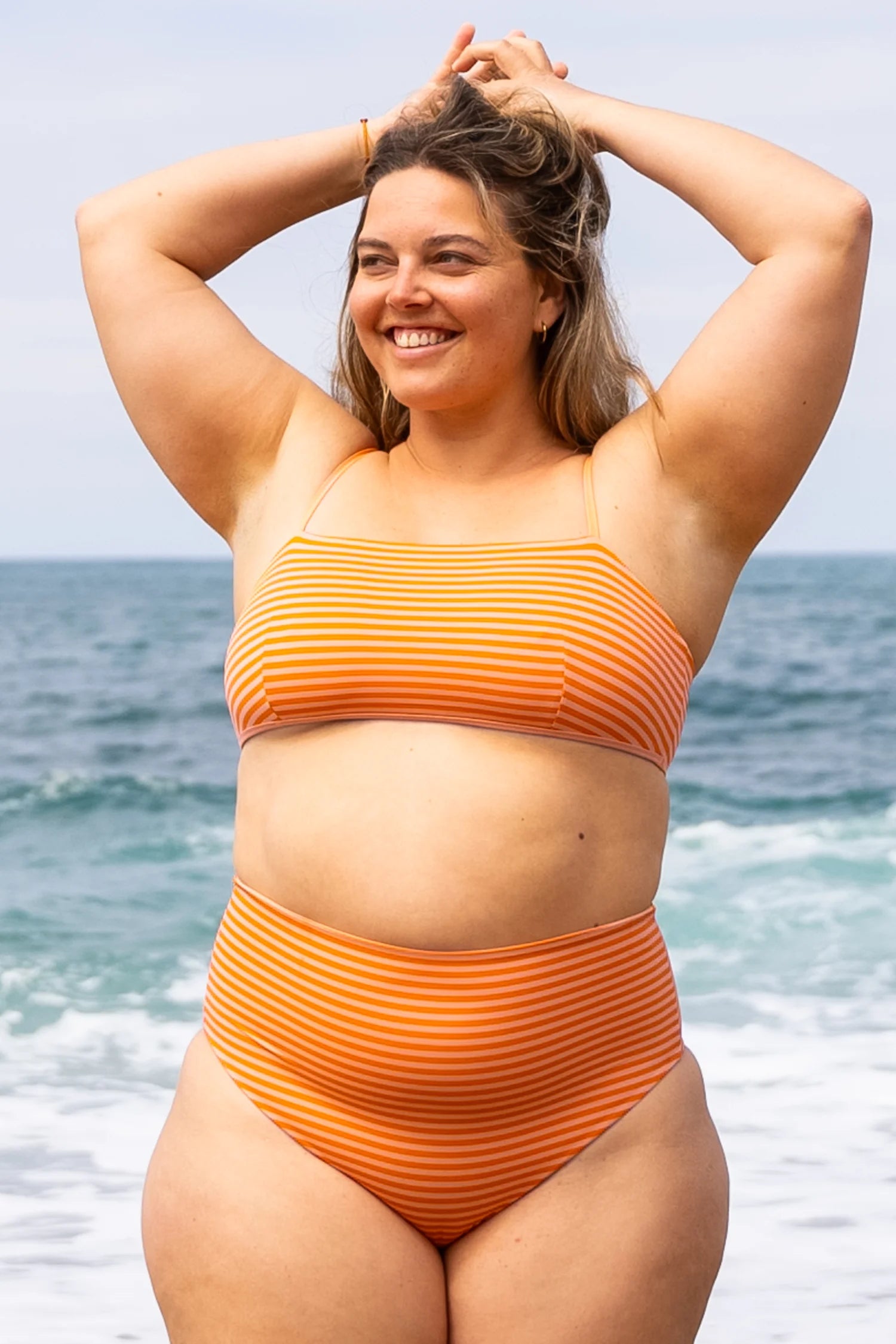 Frau am Strand trägt MYMARINI Bikini mit Spaghettiträgern und High-Waist-Hose in orange-weiß gestreift, lächelnd vor dem Meer.#color_melon-striped