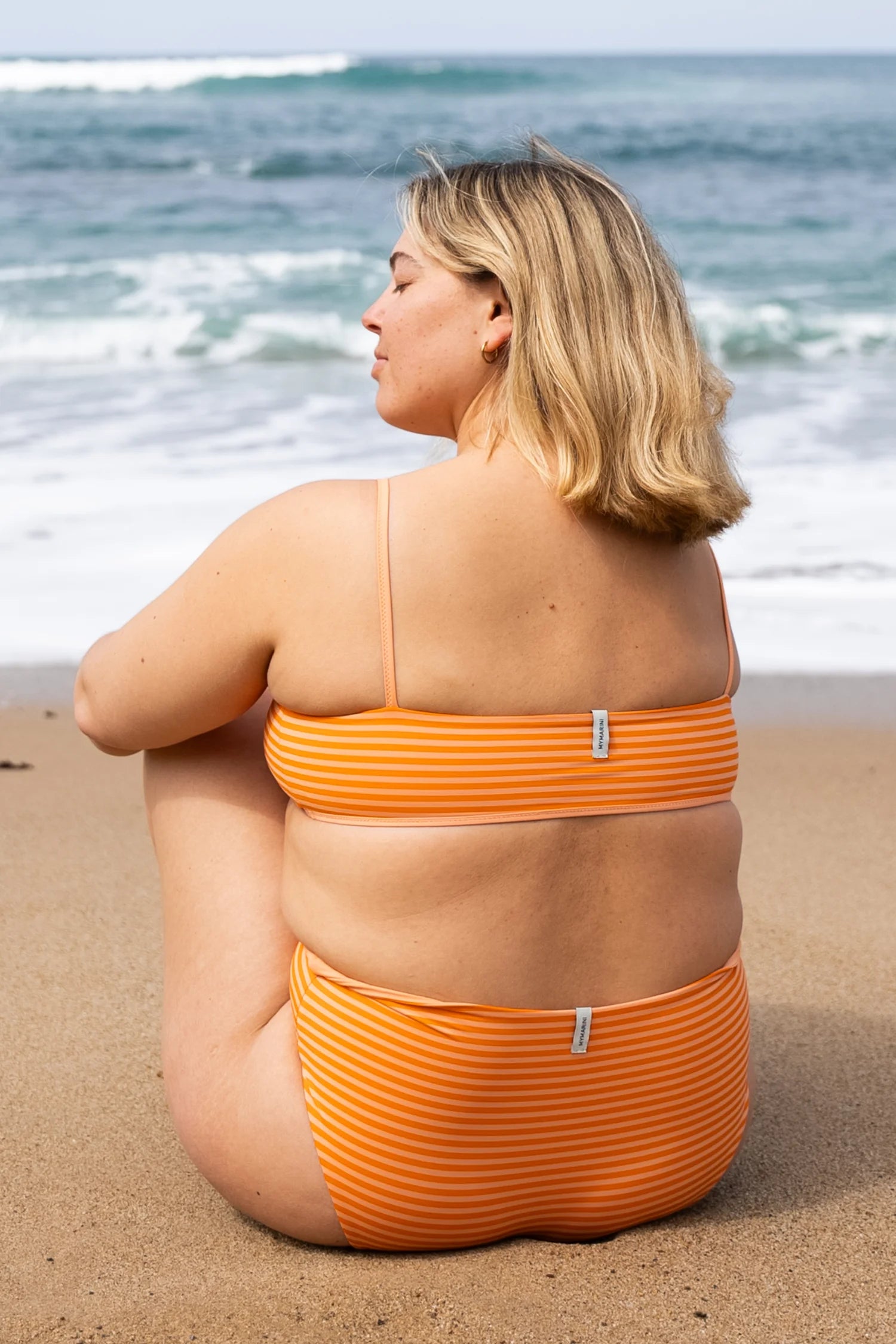 Frau am Strand in orange-gestreiftem MYMARINI Bikini mit Spaghettiträgern und hohem Beinausschnitt, blickt aufs Meer.#color_melon-striped