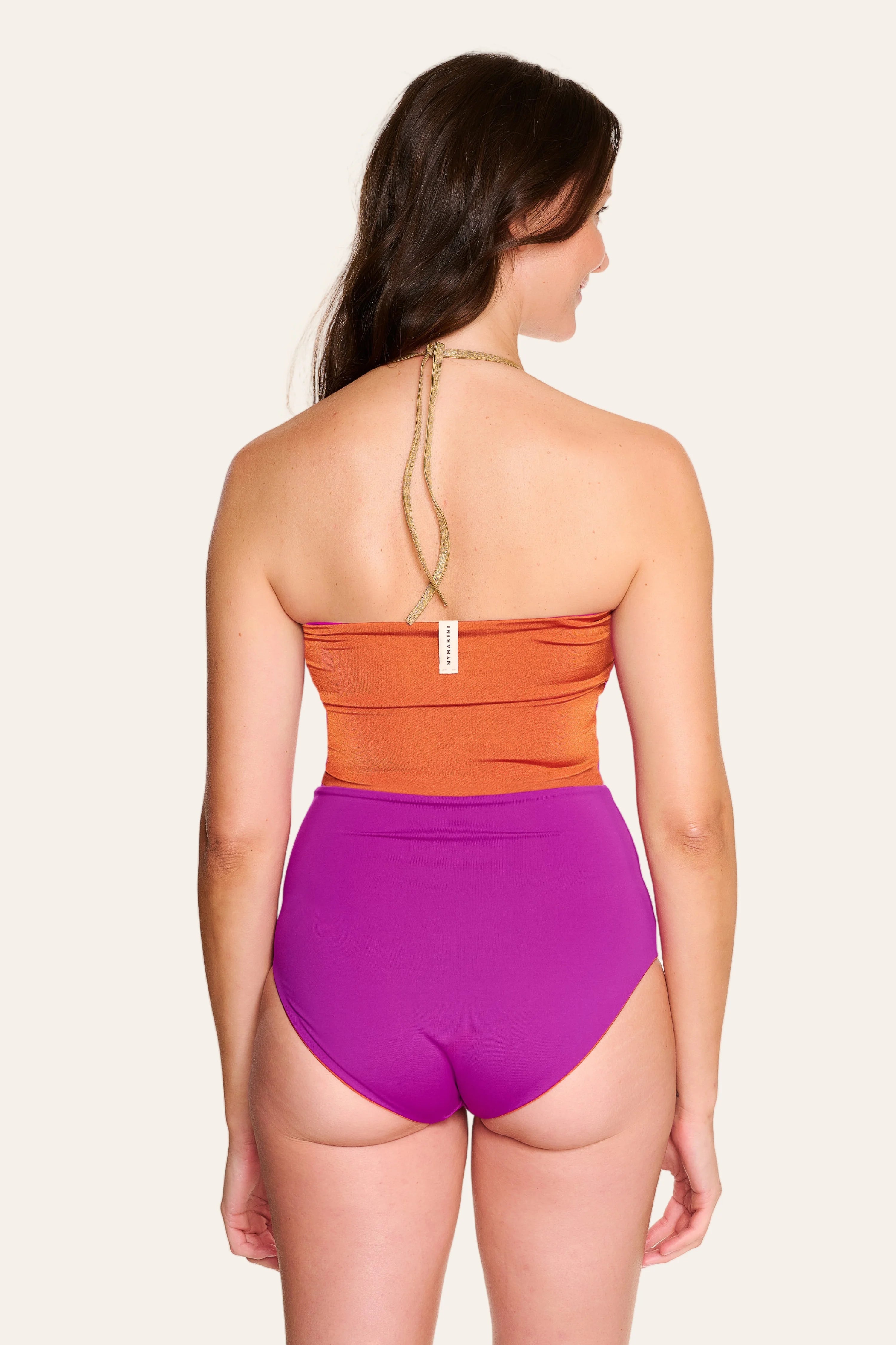 Frau trägt MYMARINI One-Piece mit Neckholder in Orange und Lila, hoher Beinausschnitt, von hinten fotografiert.#color_papaya-cassis