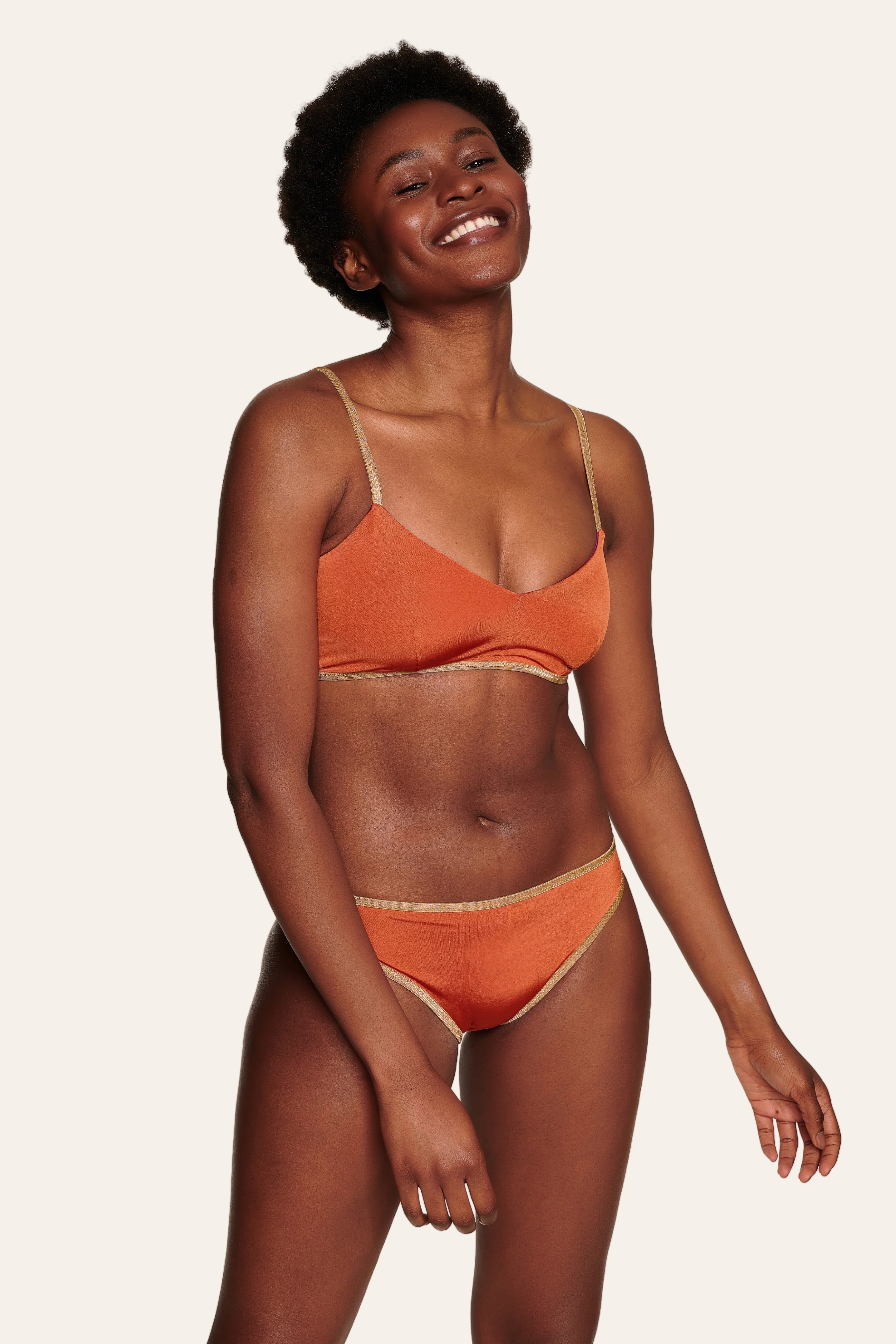 Frau trägt MYMARINI Bikini mit Spaghettiträgern und Mid rise Schnitt in Orange, lächelnd vor neutralem Hintergrund.#color_papaya-cassis