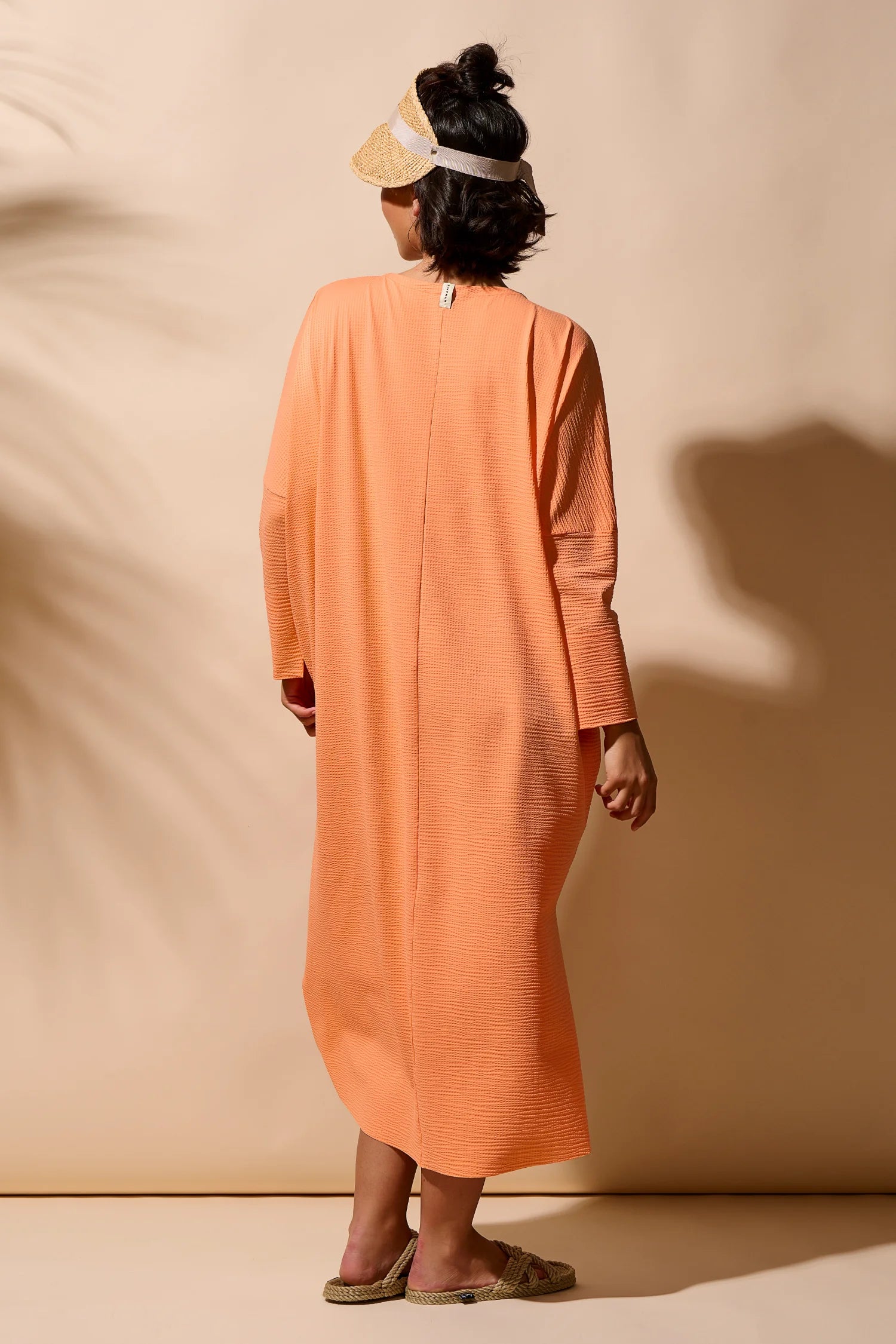 Person von hinten in einem langen, orangefarbenen MYMARINI-Kleid mit langen Ärmeln und Sandalen.#color_peach