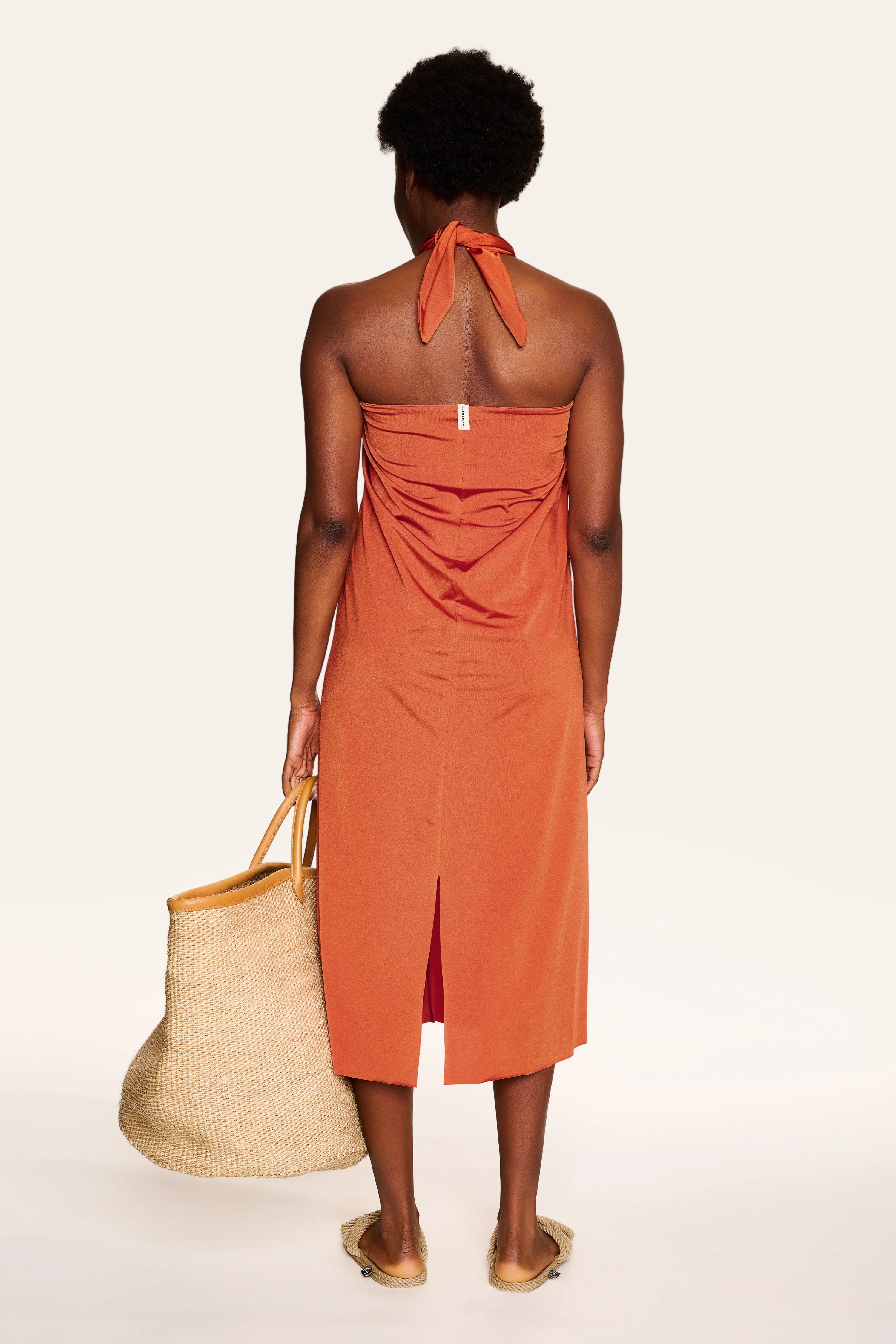 Person von hinten in einem orangefarbenen MYMARINI Neckholder-Kleid, hält eine Strohtasche.#color_papaya