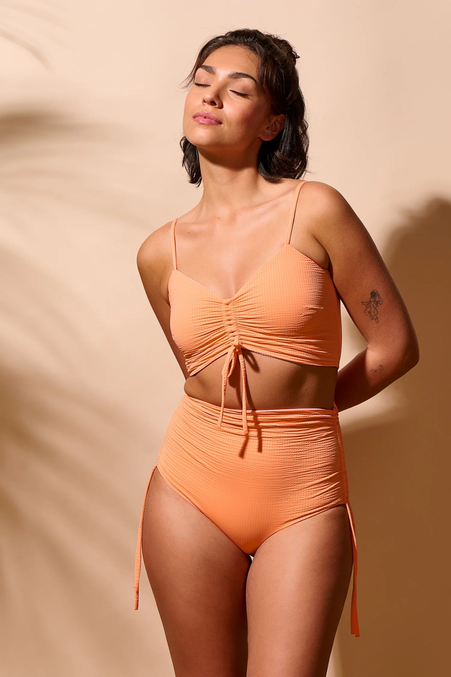Frau trägt MYMARINI Bikini mit Spaghettiträgern und High Waist in Orange vor beigem Hintergrund.#color_marshmallow-peach