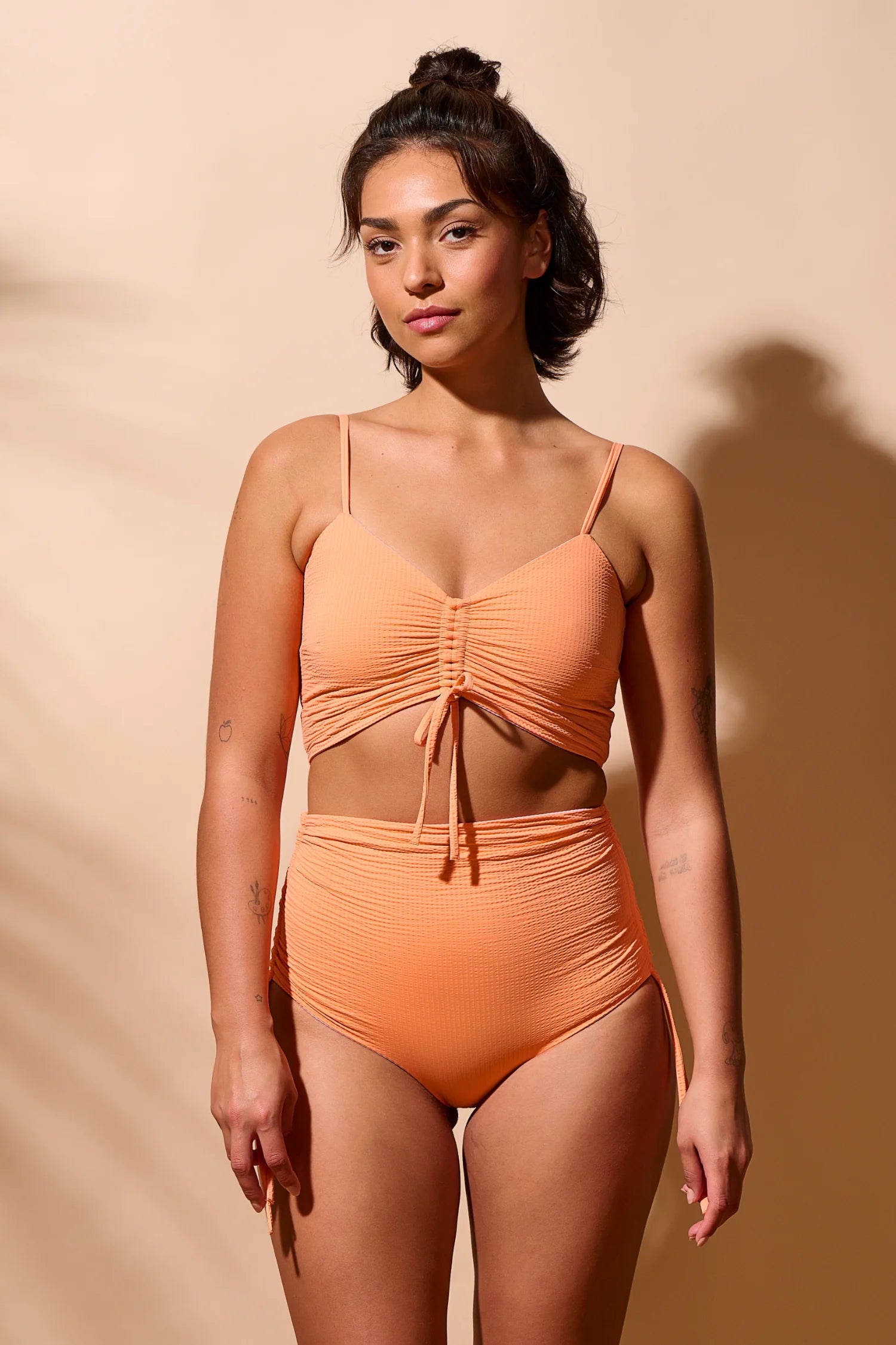 Frau trägt MYMARINI Bikini mit Spaghettiträgern und High Waist in Apricot vor beigem Hintergrund.#color_marshmallow-peach